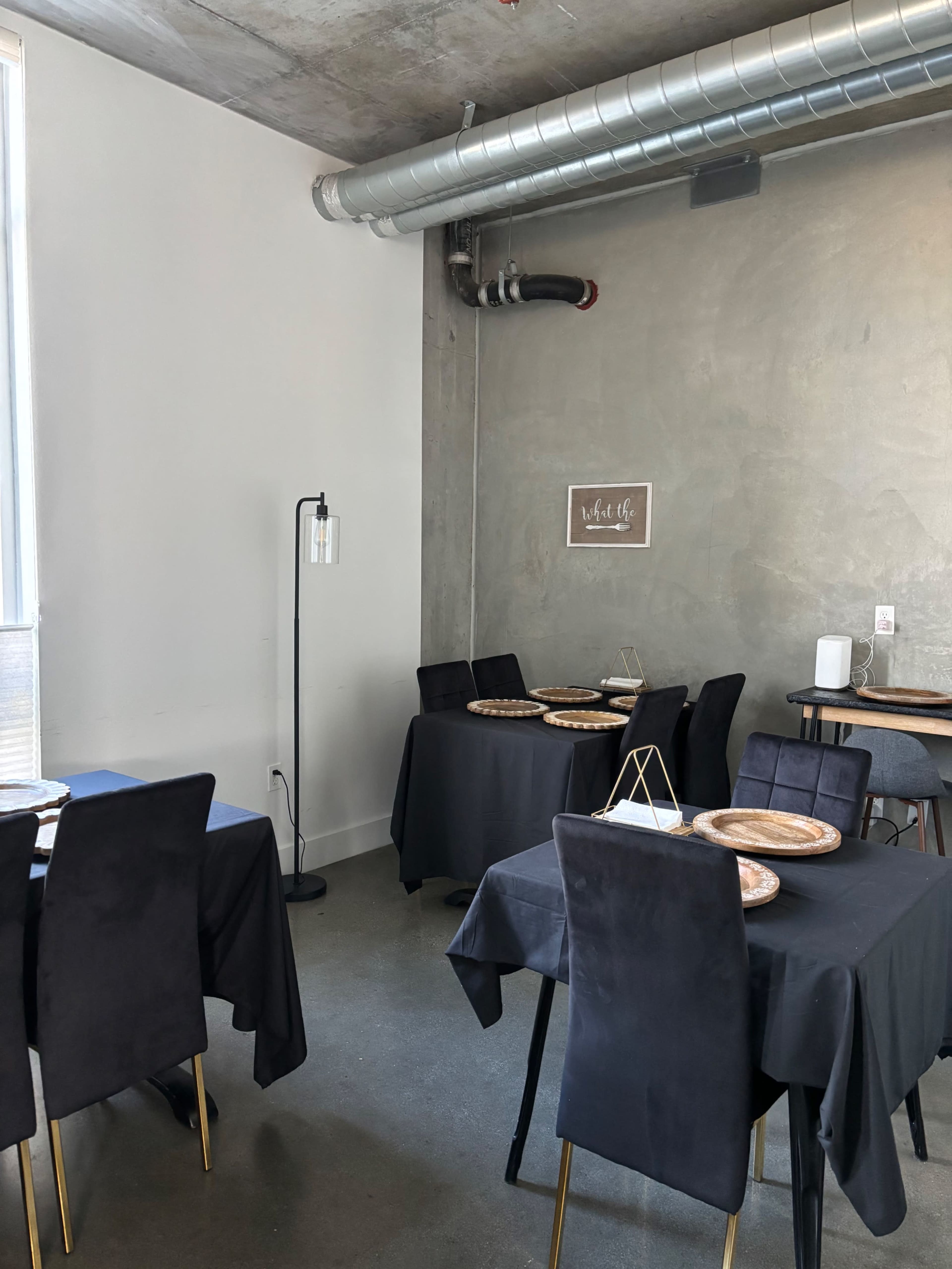 The image shows a minimalist dining area with black tables covered in dark tablecloths and wooden serving platters, complemented by modern seating and a concrete wall.
