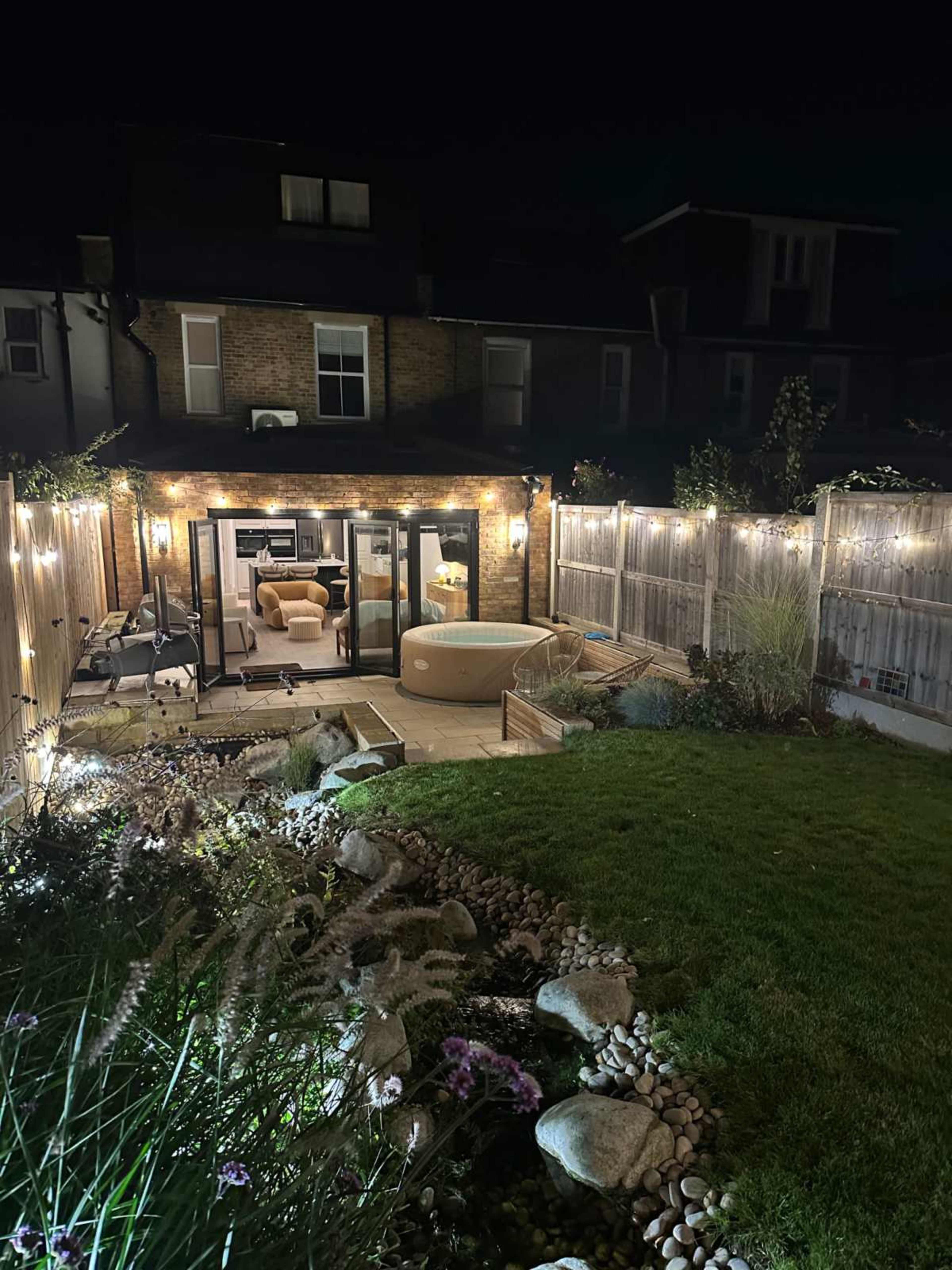 The image shows a well-lit backyard at night, featuring a hot tub, landscaped stone pathways, and modern outdoor furniture.