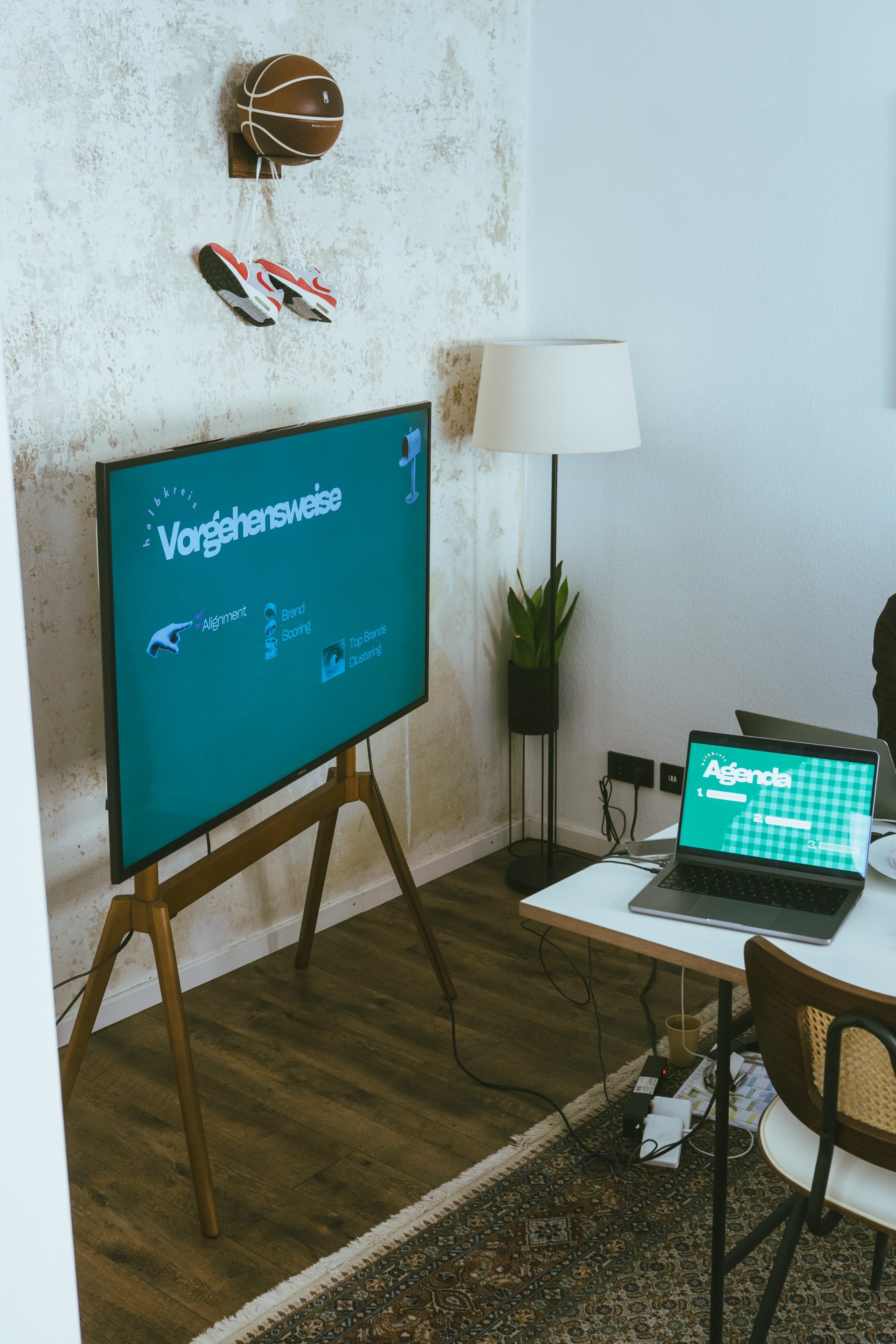 A large television displays a presentation titled "Vorgehensweise" while a laptop sits on a table with various cables visible, and a lamp and sports items are positioned in the background.