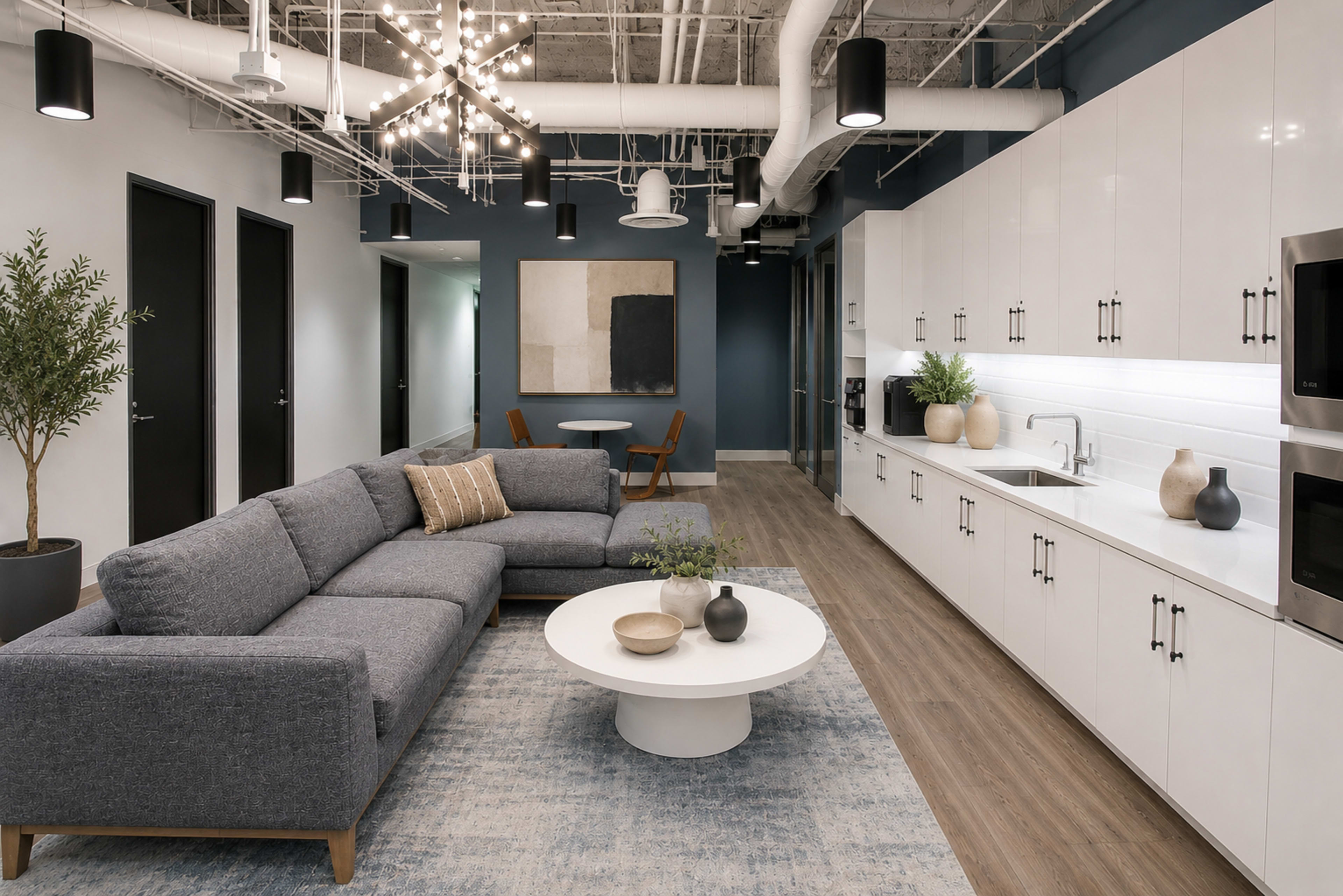 The image shows a modern break room with a gray sectional sofa, a round coffee table, and a kitchenette featuring white cabinets and stainless steel appliances.