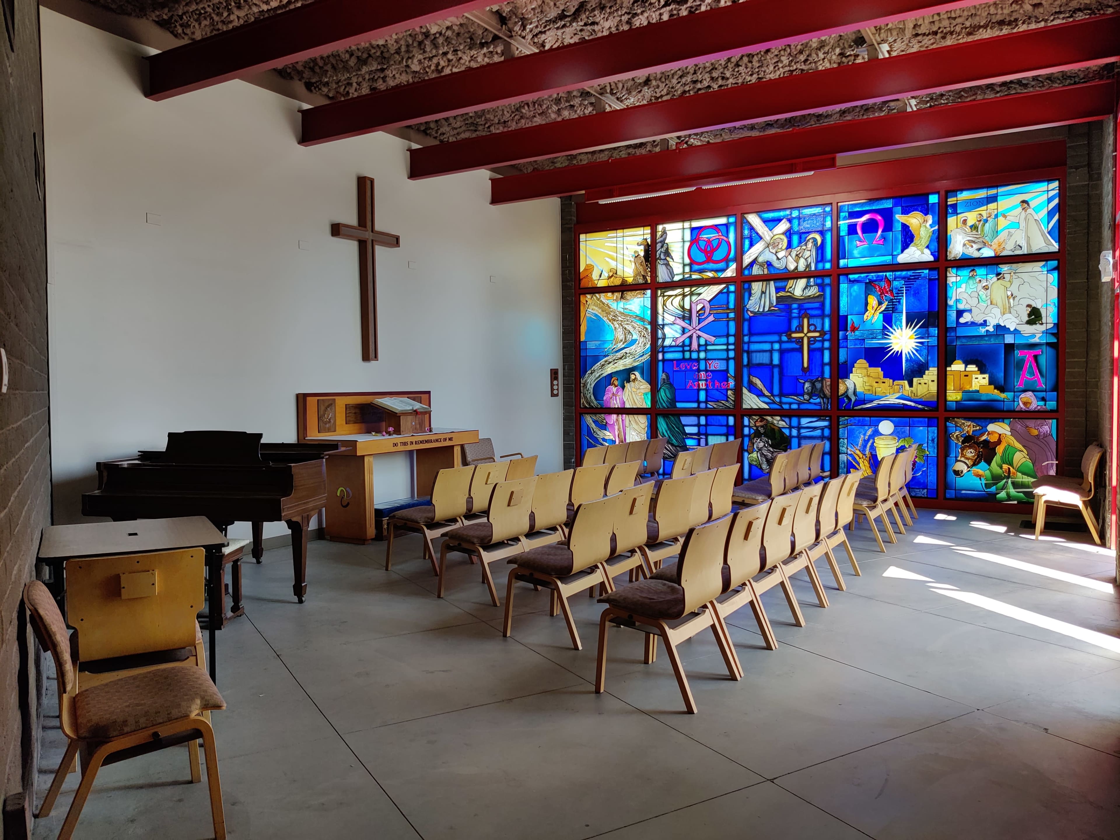 A brightly lit room featuring a cross on one wall, a piano, wooden furniture, and rows of empty chairs facing a large stained glass window.