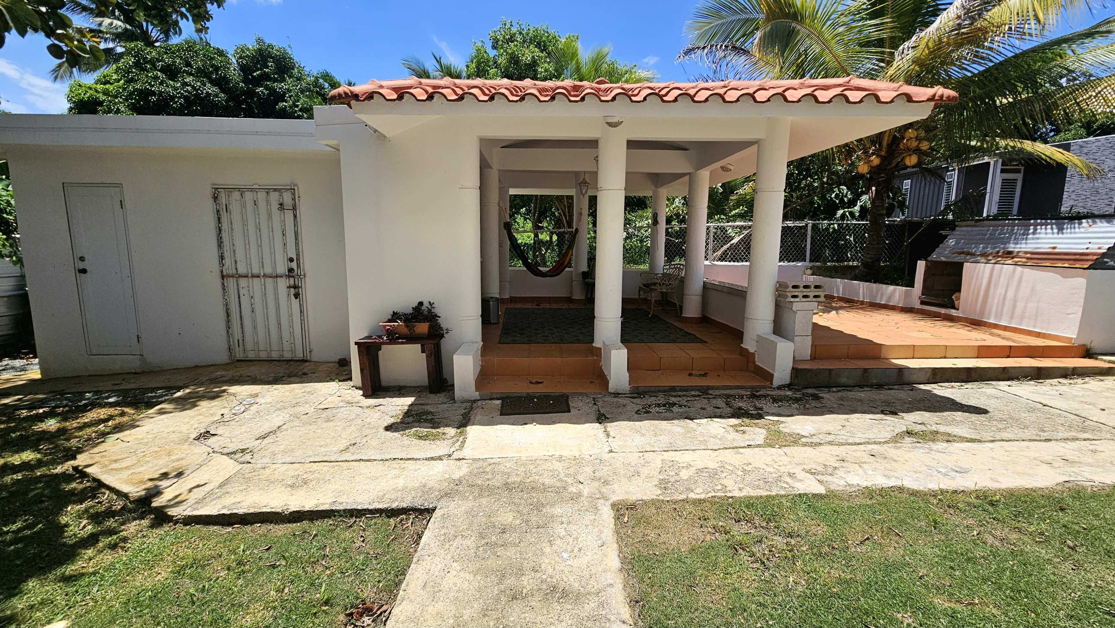 The image shows a house with a tiled porch, supported by columns, a hammock hanging on one side, and a grassy area in front.
