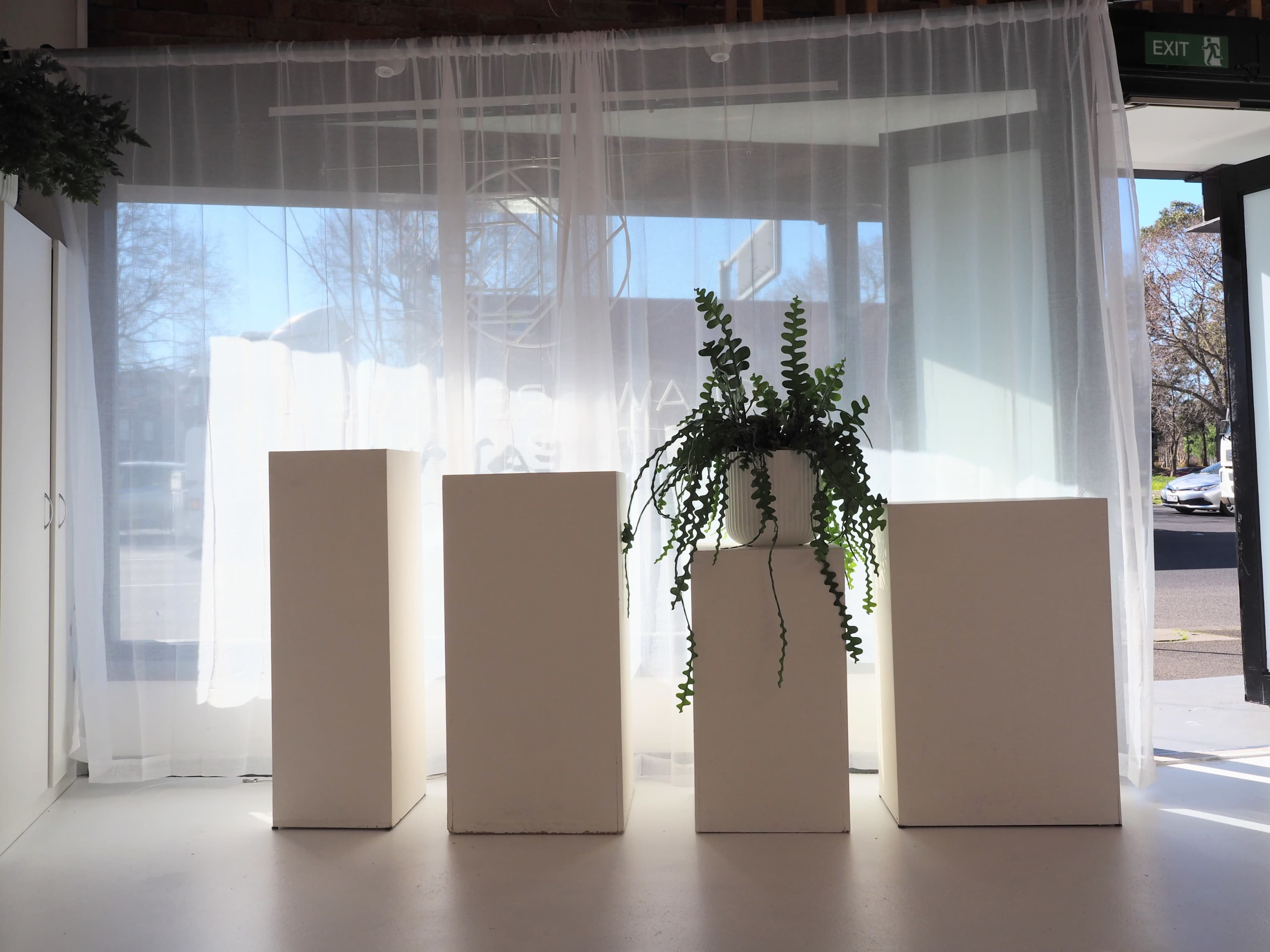 The image shows four white pedestals of varying heights placed in front of a sheer curtain and a large window.