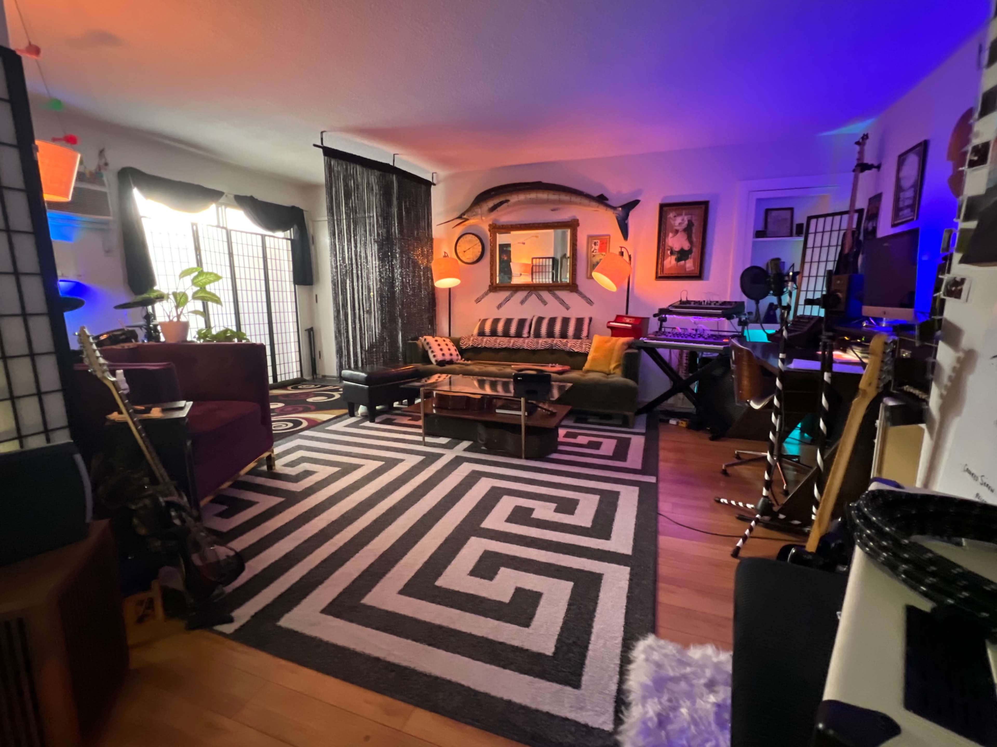 The room features a patterned black and white rug, various seating areas, plants, and a mix of musical instruments and electronic equipment.