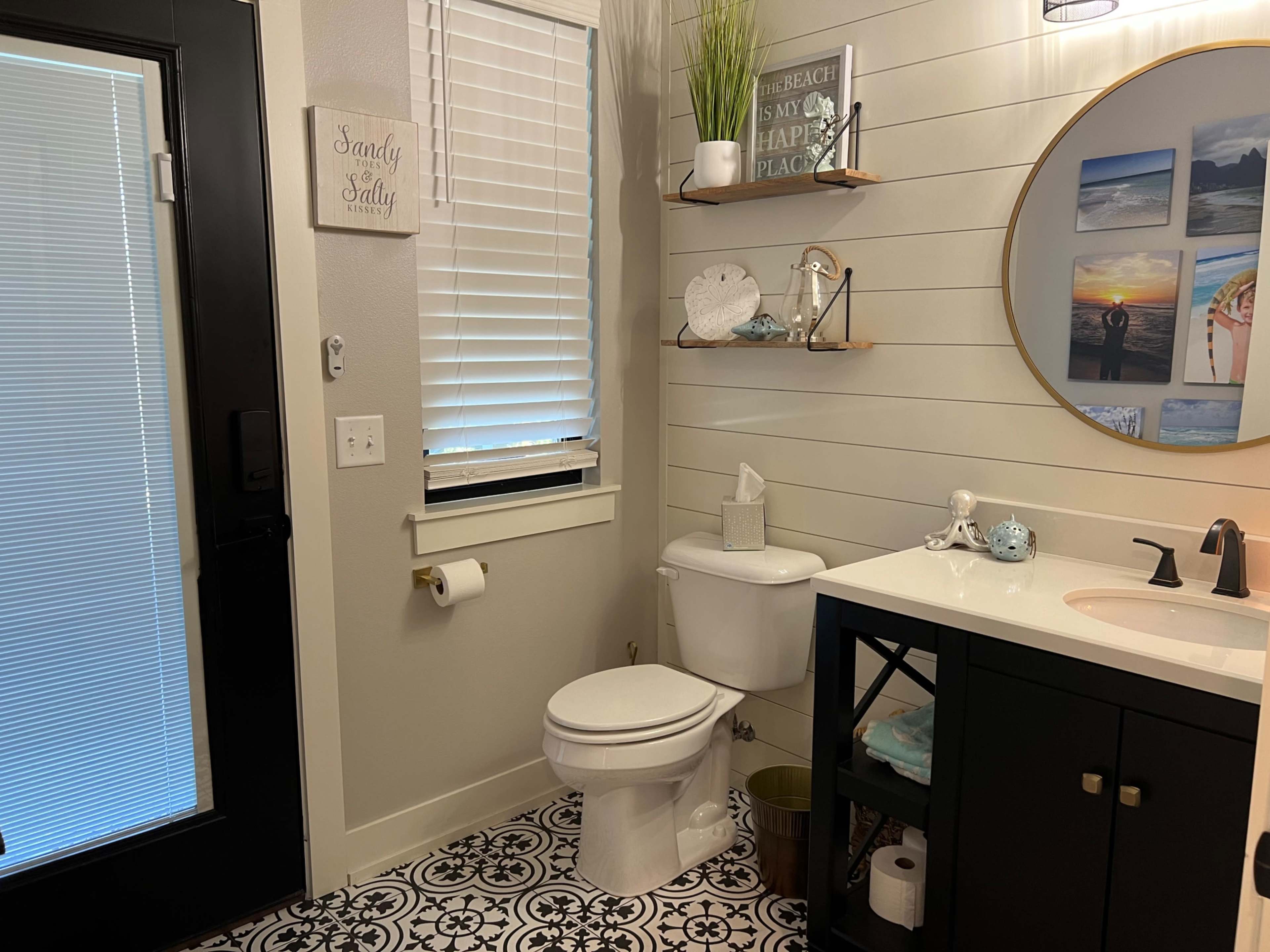 The image shows a modern bathroom featuring a white toilet, a dark vanity with open shelving, and a round mirror above the sink.