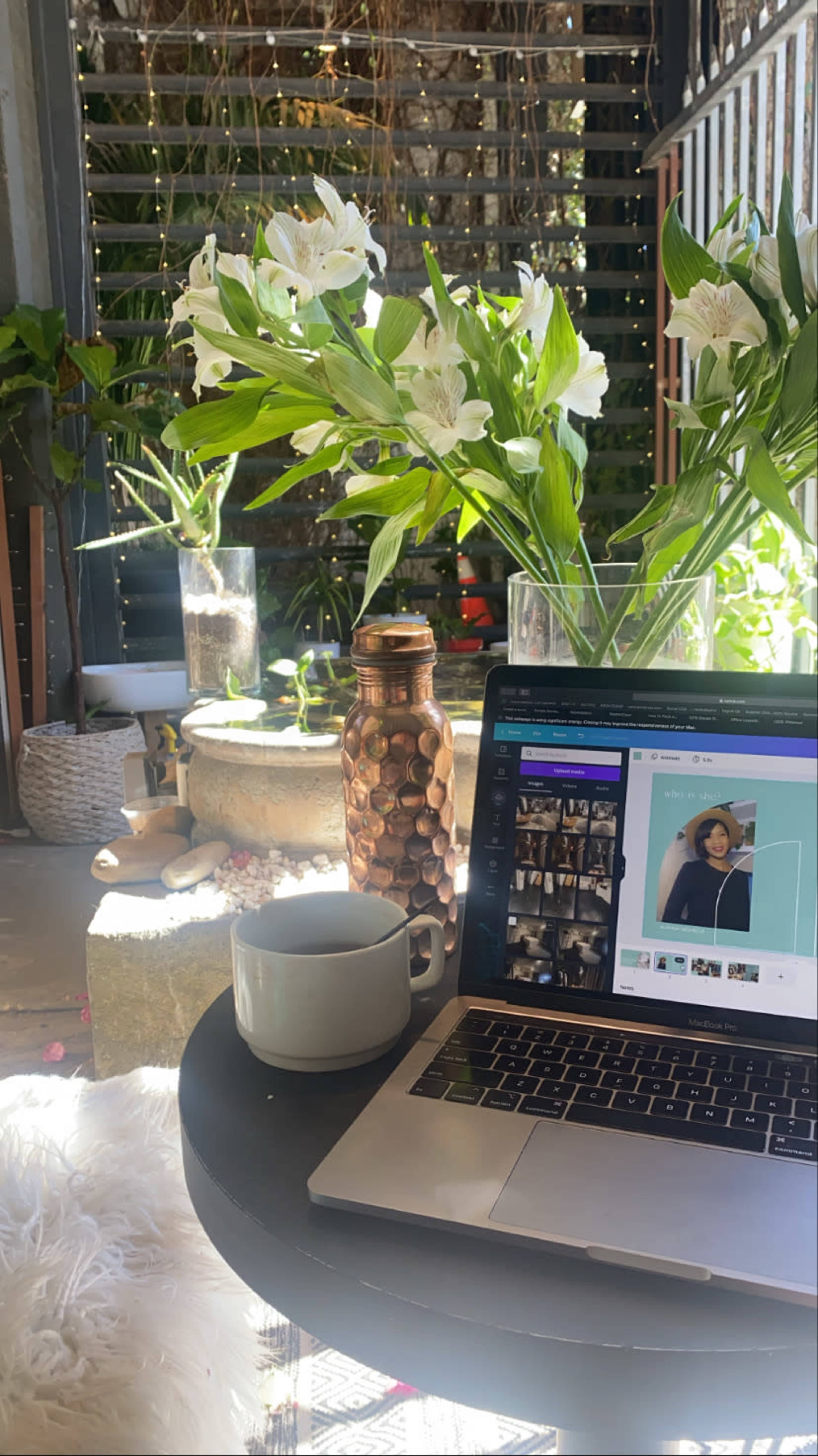 A laptop displays a design interface on a table beside a cup and a large vase of flowers in a sunlit outdoor space.