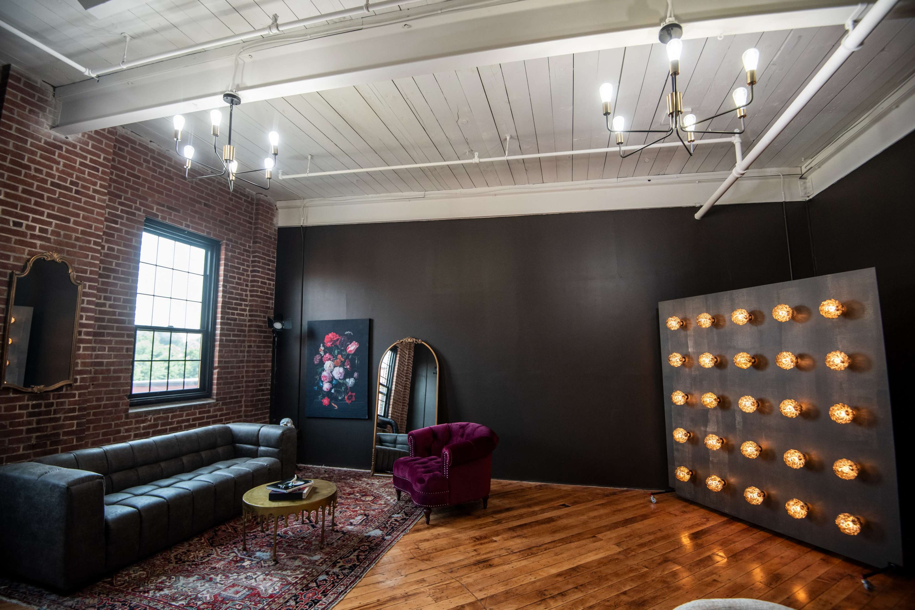 A spacious living area featuring a gray sofa, a round table, a purple chair, and a decorative wall with lights, against a backdrop of exposed brick and dark walls.