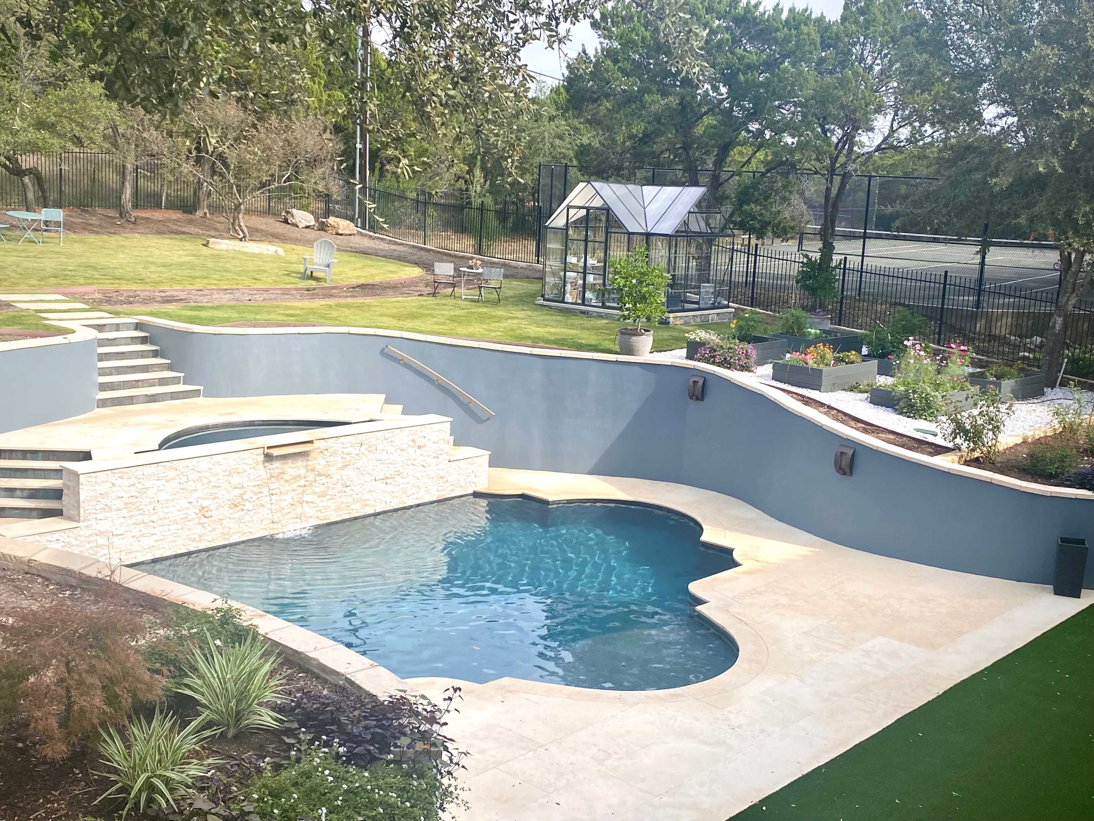 The image shows a backyard with a swimming pool featuring a stone wall, landscaped greenery, and a greenhouse in the background.