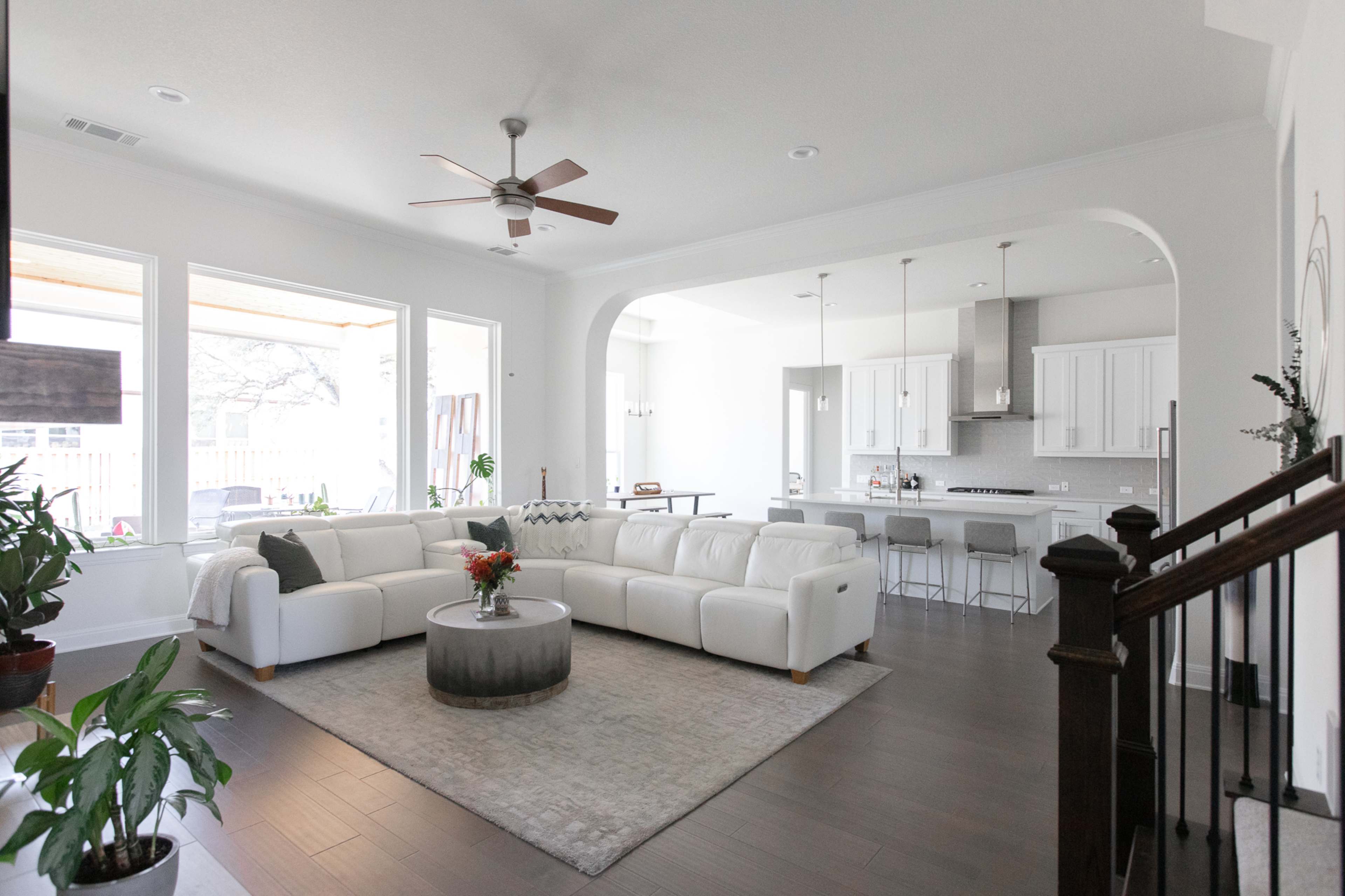 A spacious living room features a white sectional sofa, a round coffee table, and a view of an adjacent kitchen with modern appliances and fixtures.