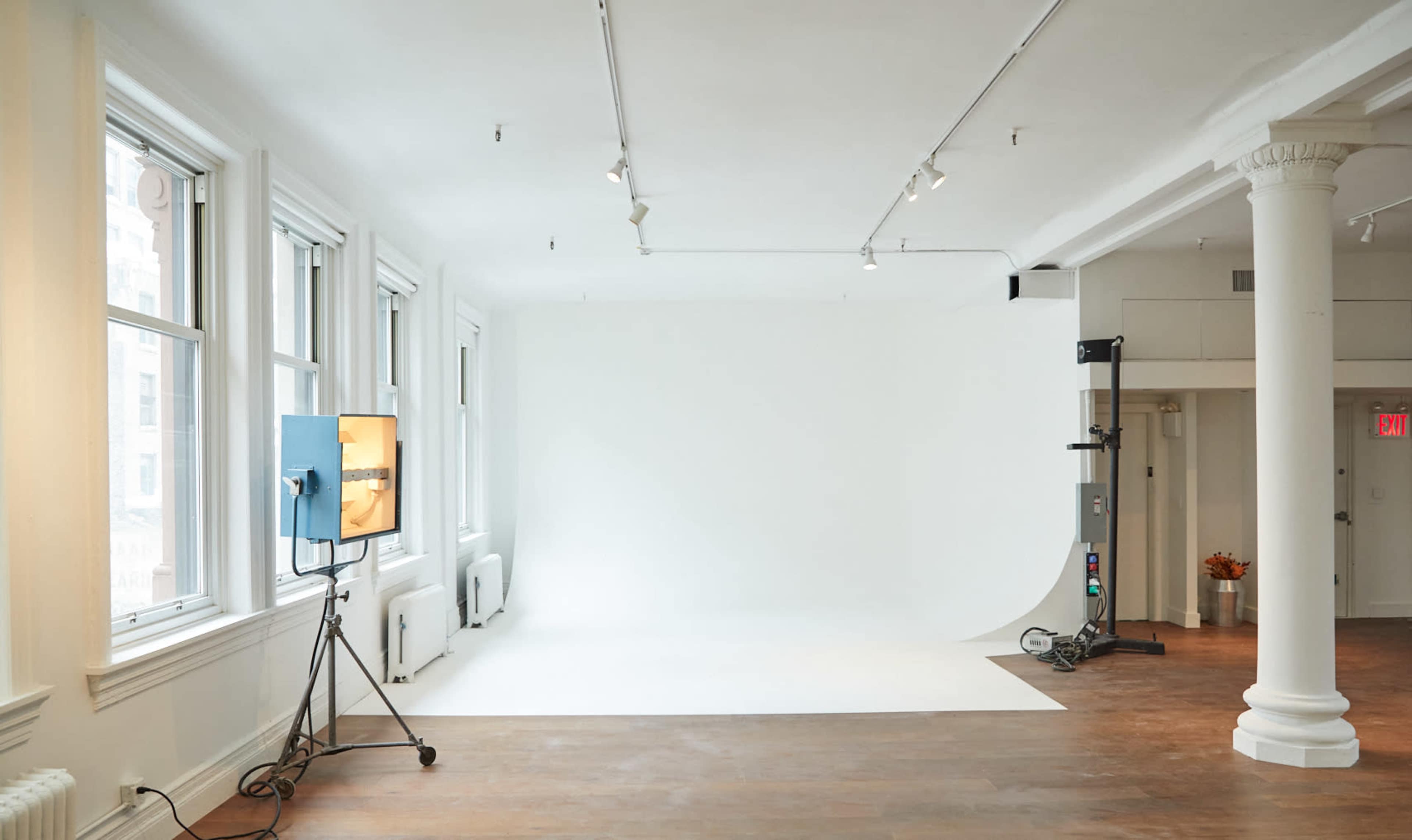 The image shows a spacious photography studio with large windows, a white backdrop, and minimal furnishings.