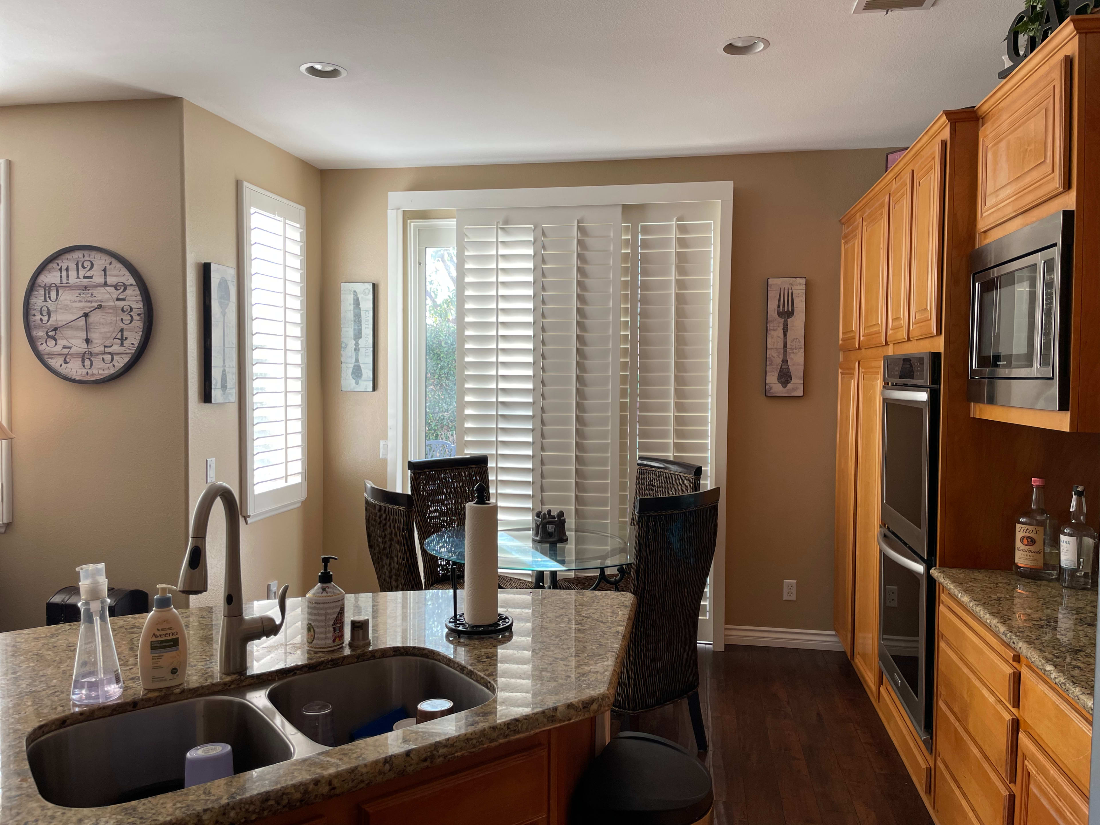 A well-lit kitchen with wooden cabinets, a granite countertop, a circular dining table with chairs, and a window covered with white shutters.