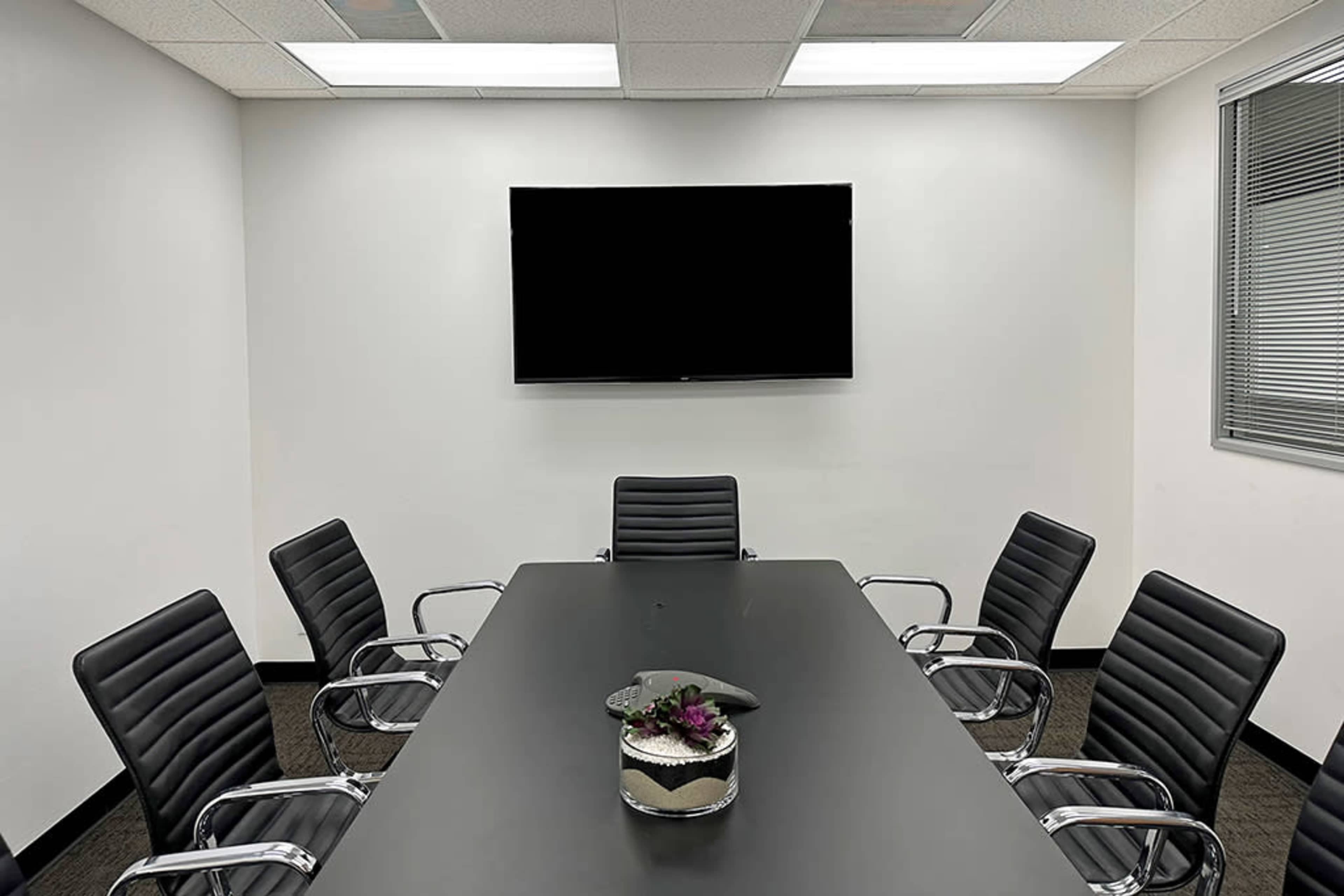 A conference room features a long black table surrounded by several black leather chairs and a large wall-mounted television.