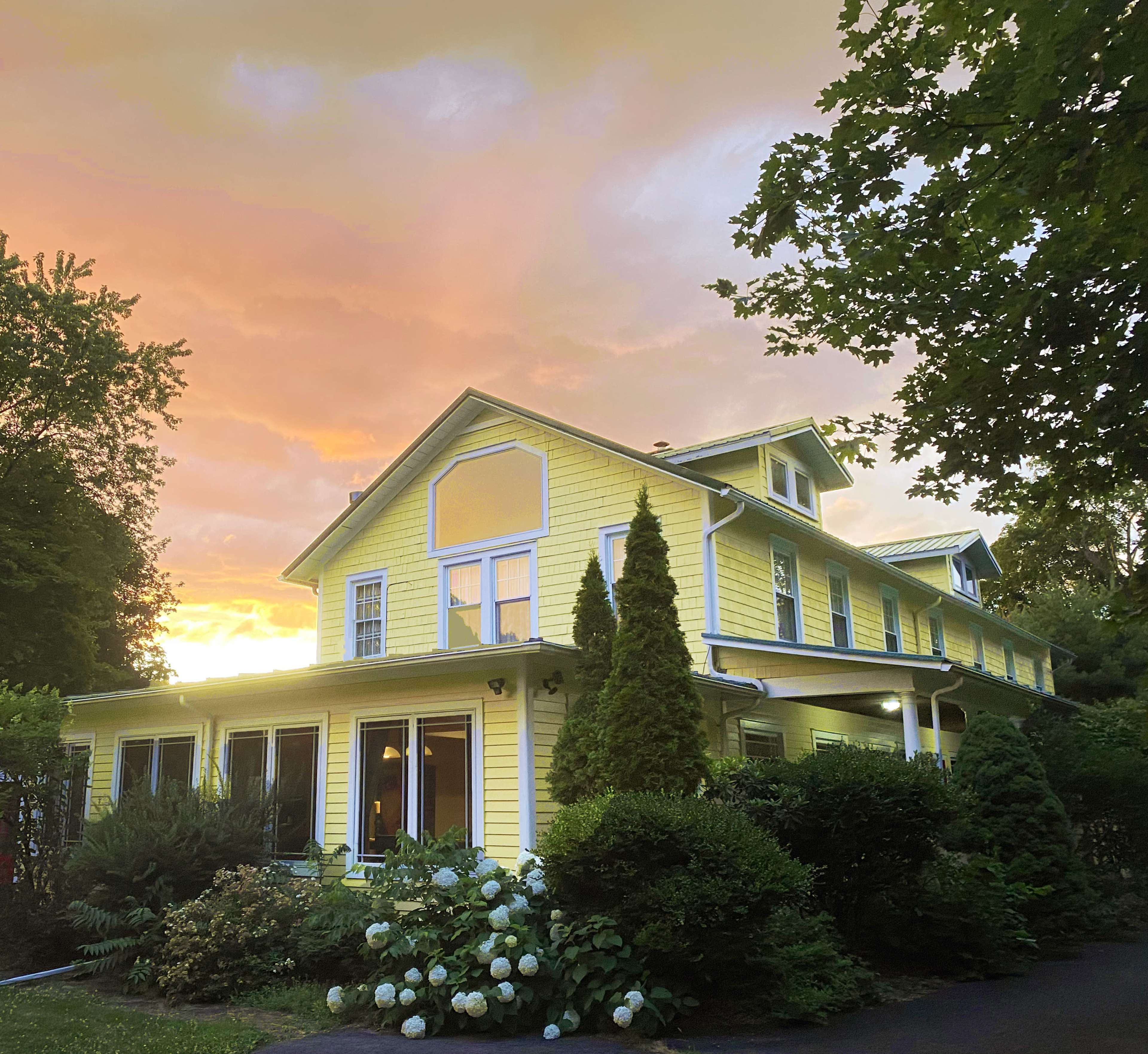 A yellow house with multiple windows is framed by greenery, under a sky illuminated by a colorful sunset.