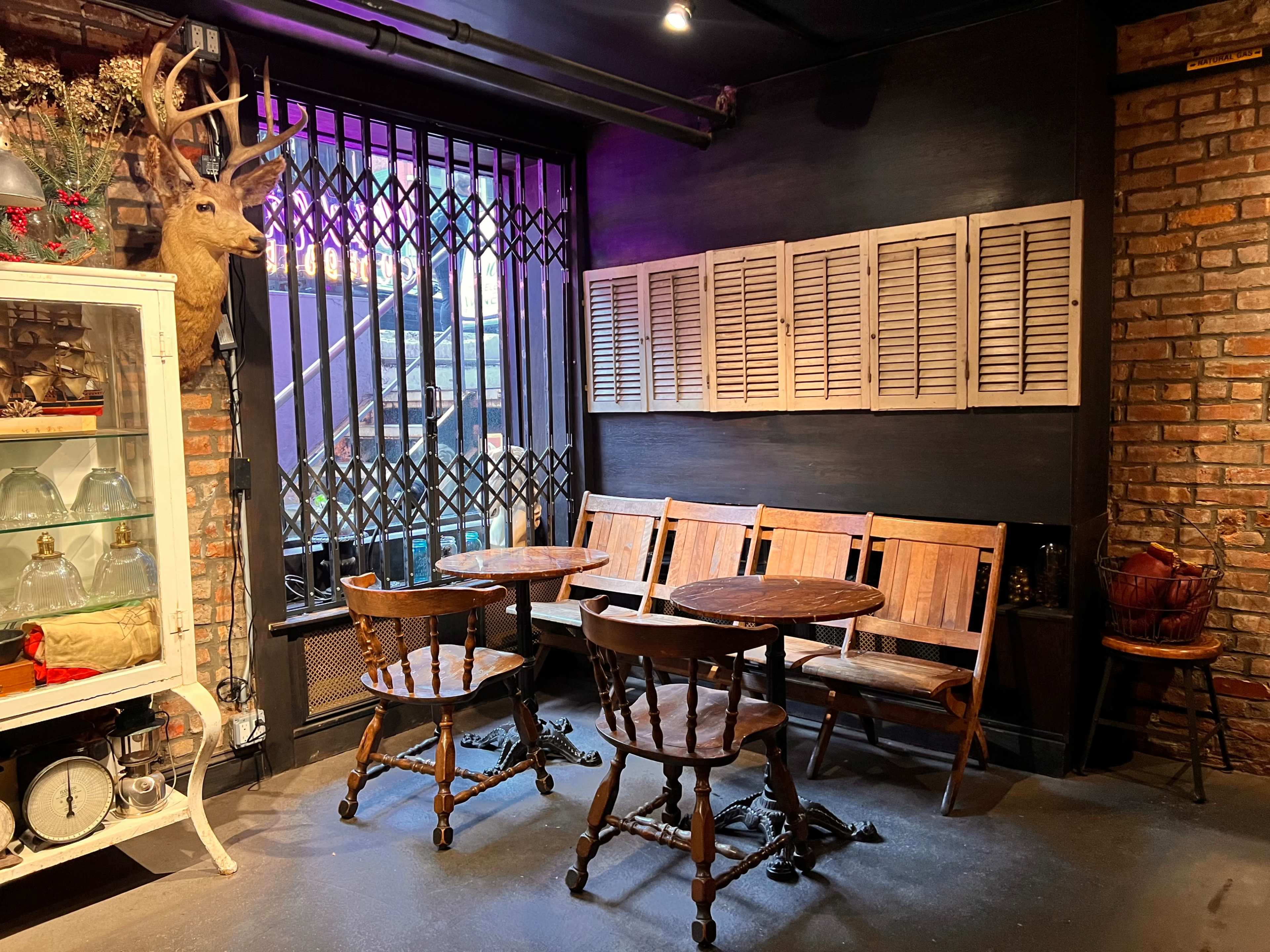The image shows a cozy seating area with wooden tables and chairs, set against a backdrop of exposed brick walls and decorative elements, including a taxidermy deer head and a glass cabinet.