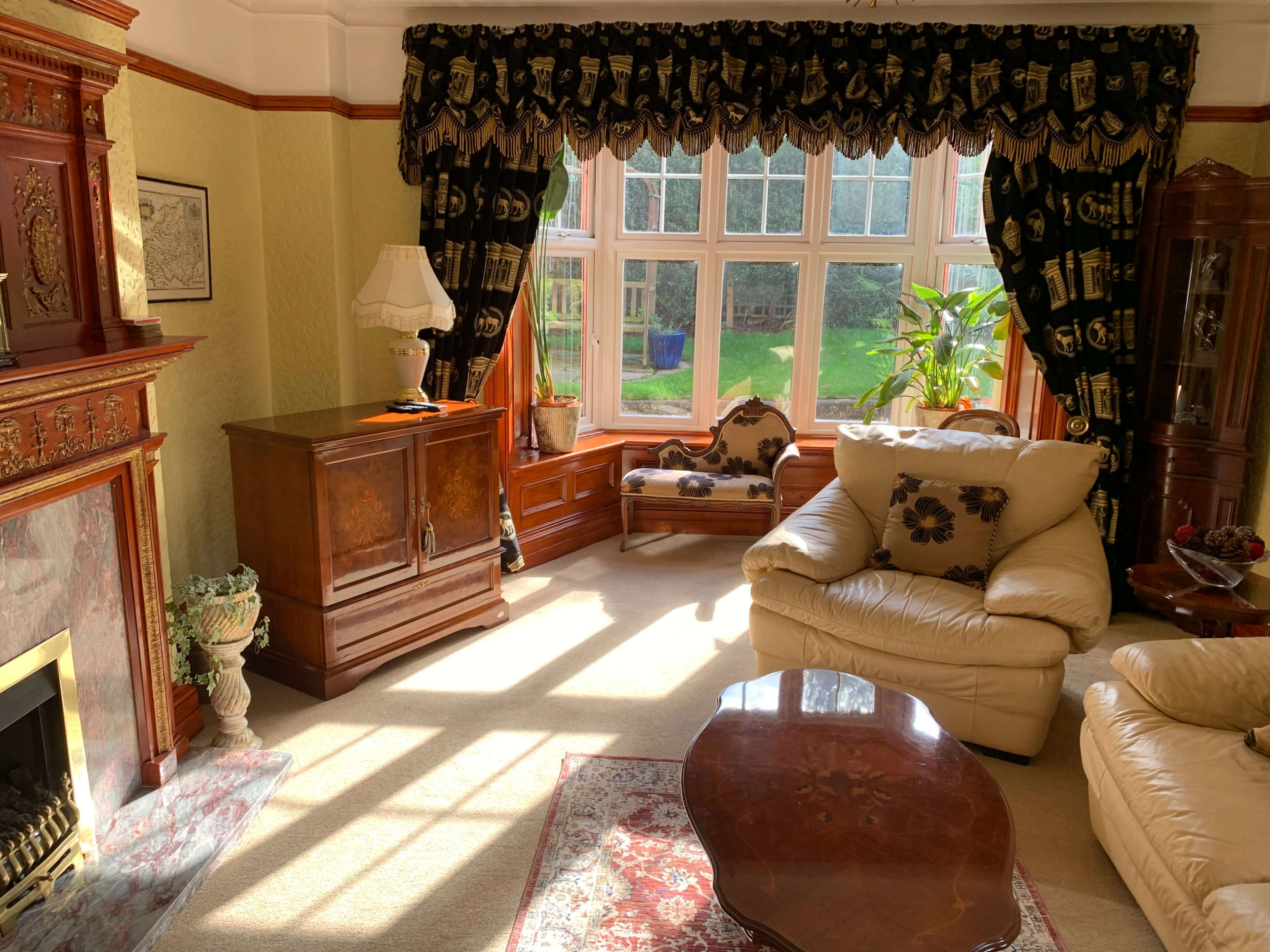 The living room features a large window with drapes, a fireplace, and a mix of wooden furniture and upholstered seating.