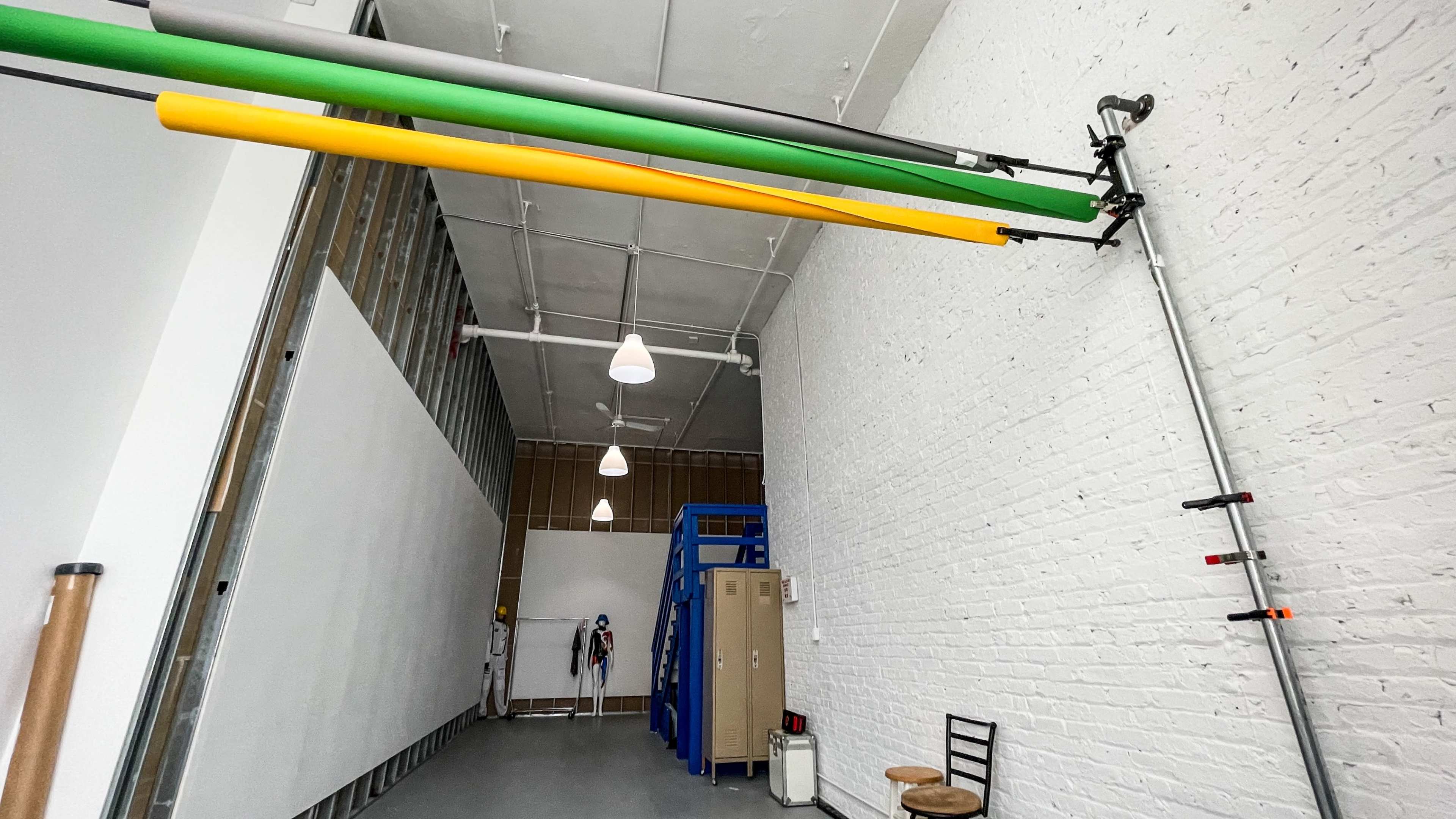 The image shows a spacious studio with white brick walls, overhead pipe rails in yellow and green, and a large white backdrop against one side.