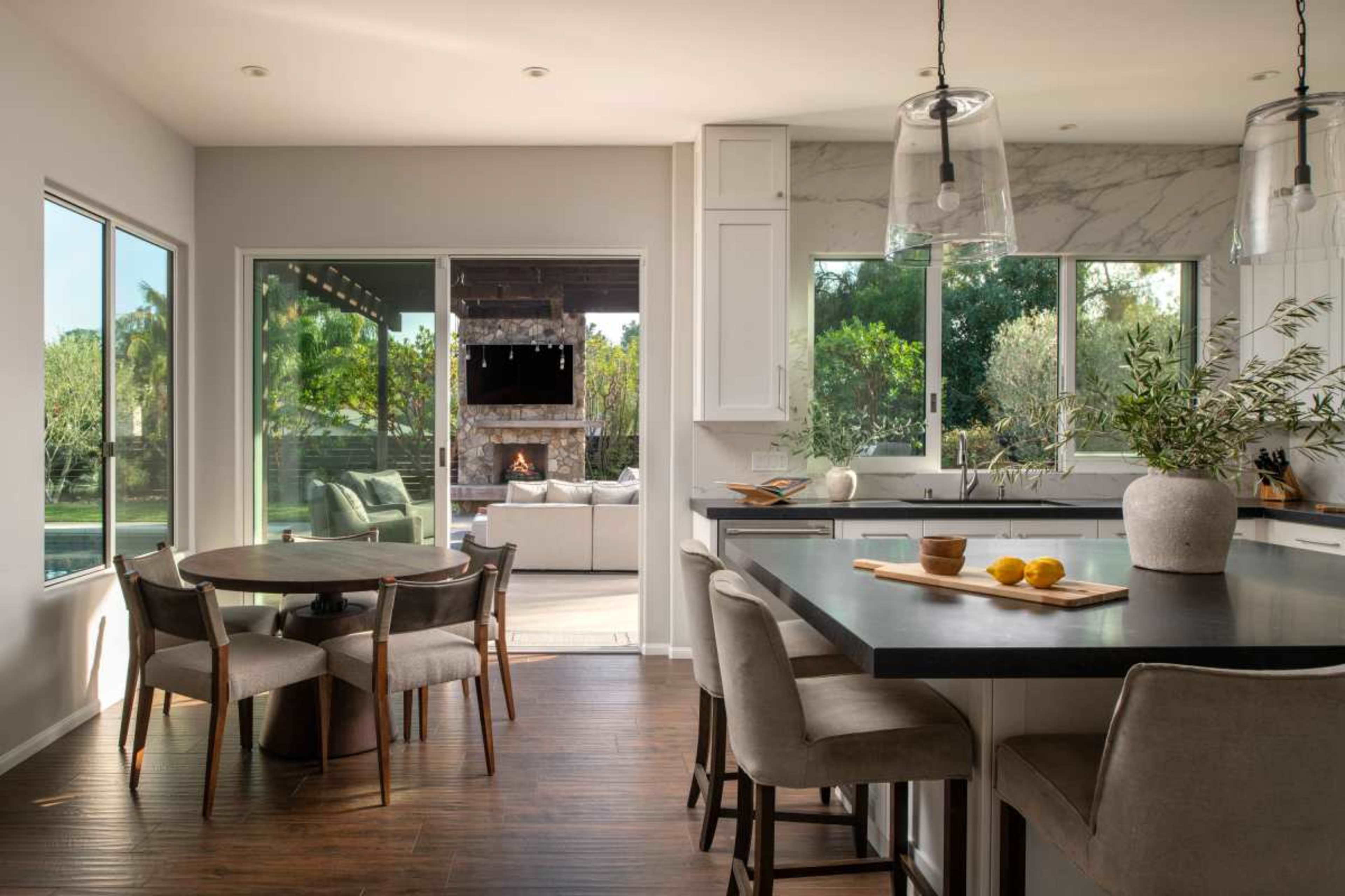 A modern kitchen features a dining area with a round table and four chairs, adjacent to large windows that overlook an outdoor seating space with a fireplace.