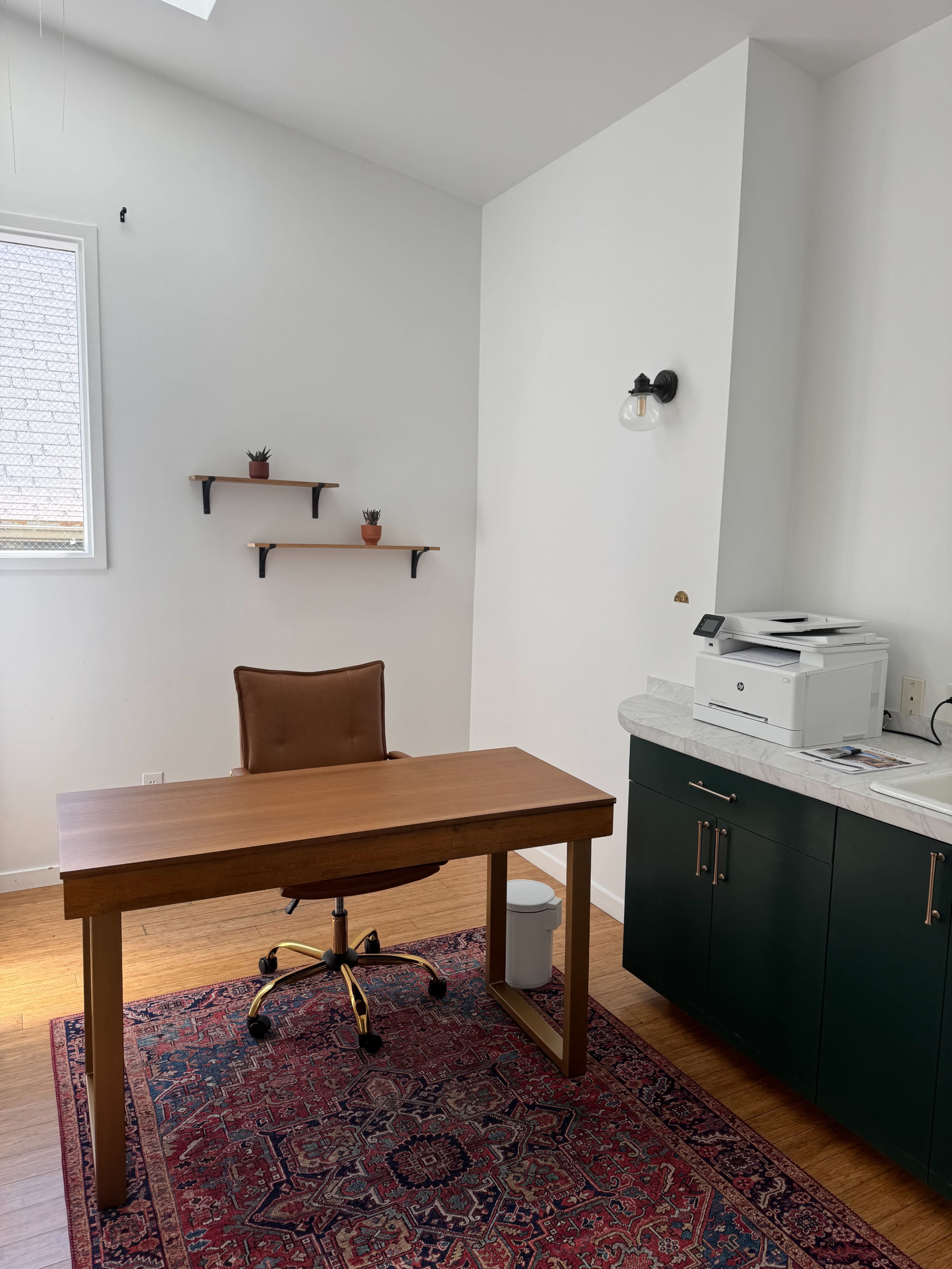The image shows a minimalist office with a wooden desk, a leather chair, a printer, and shelves with small plants on the walls.