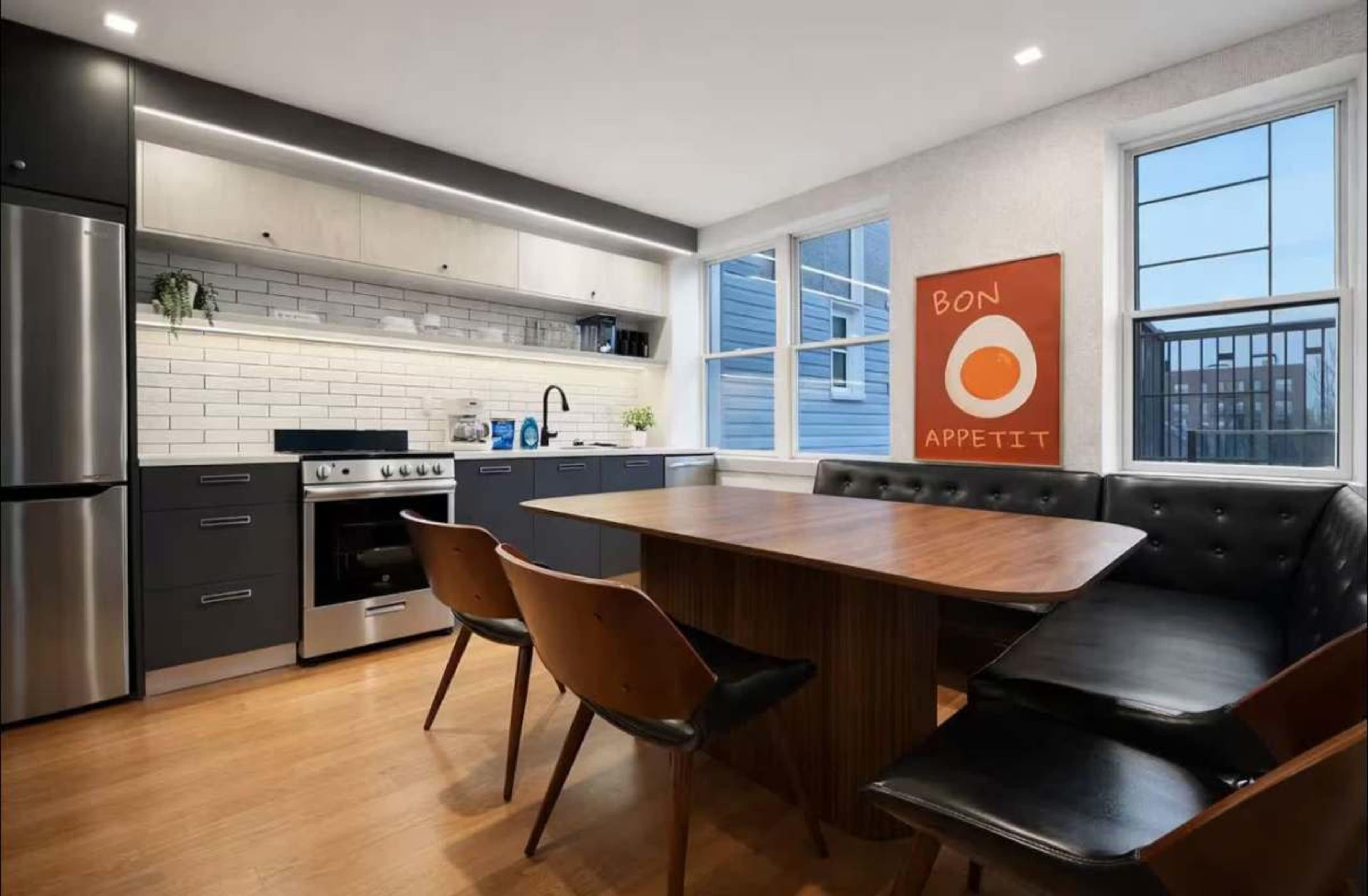 The image shows a modern kitchen featuring a stainless steel refrigerator, a stove with an oven, and a wooden dining table surrounded by black chairs.
