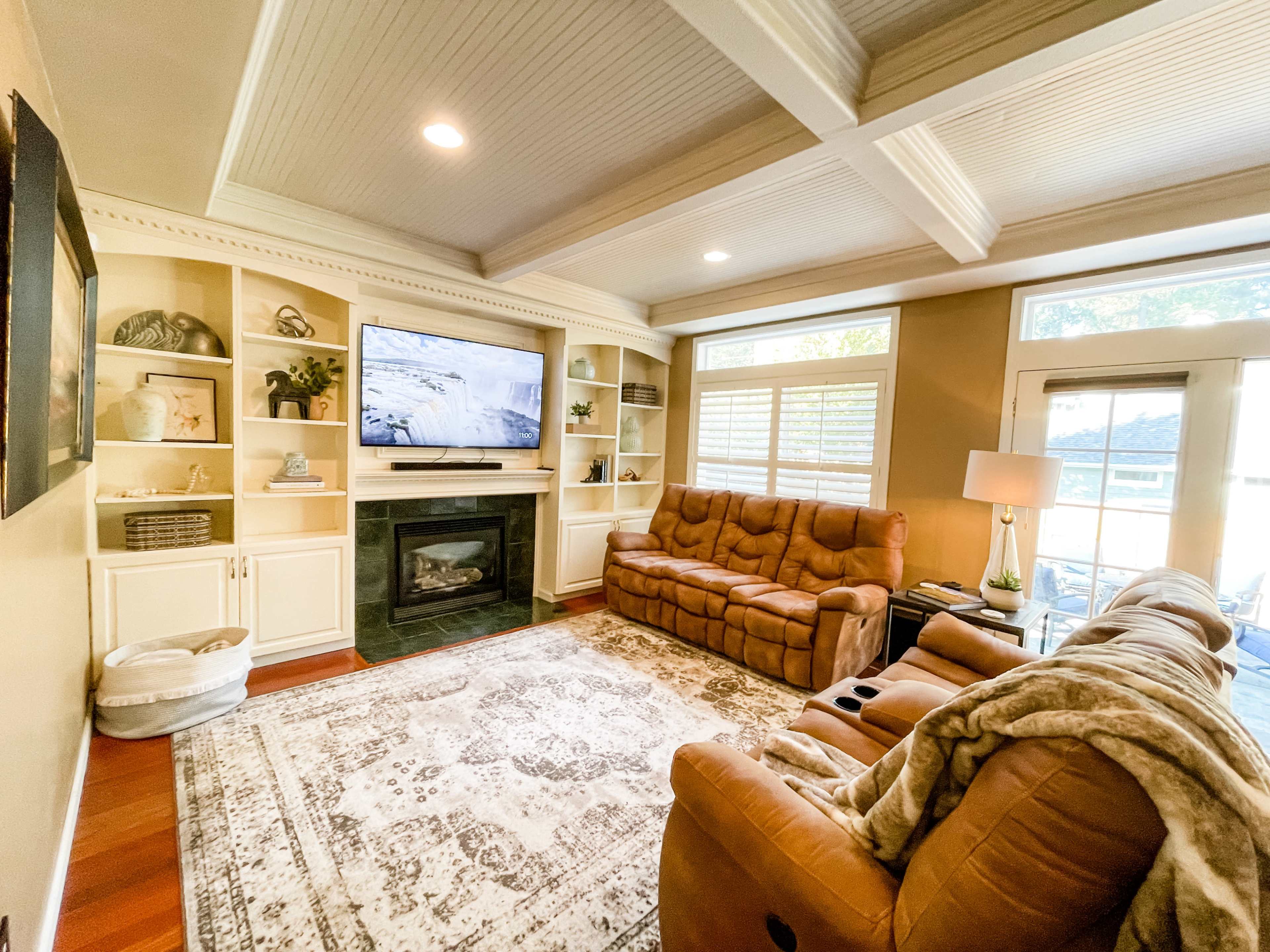 A cozy living room with a brown reclining sofa, a television mounted above a black fireplace, built-in shelves, and large windows allowing natural light.