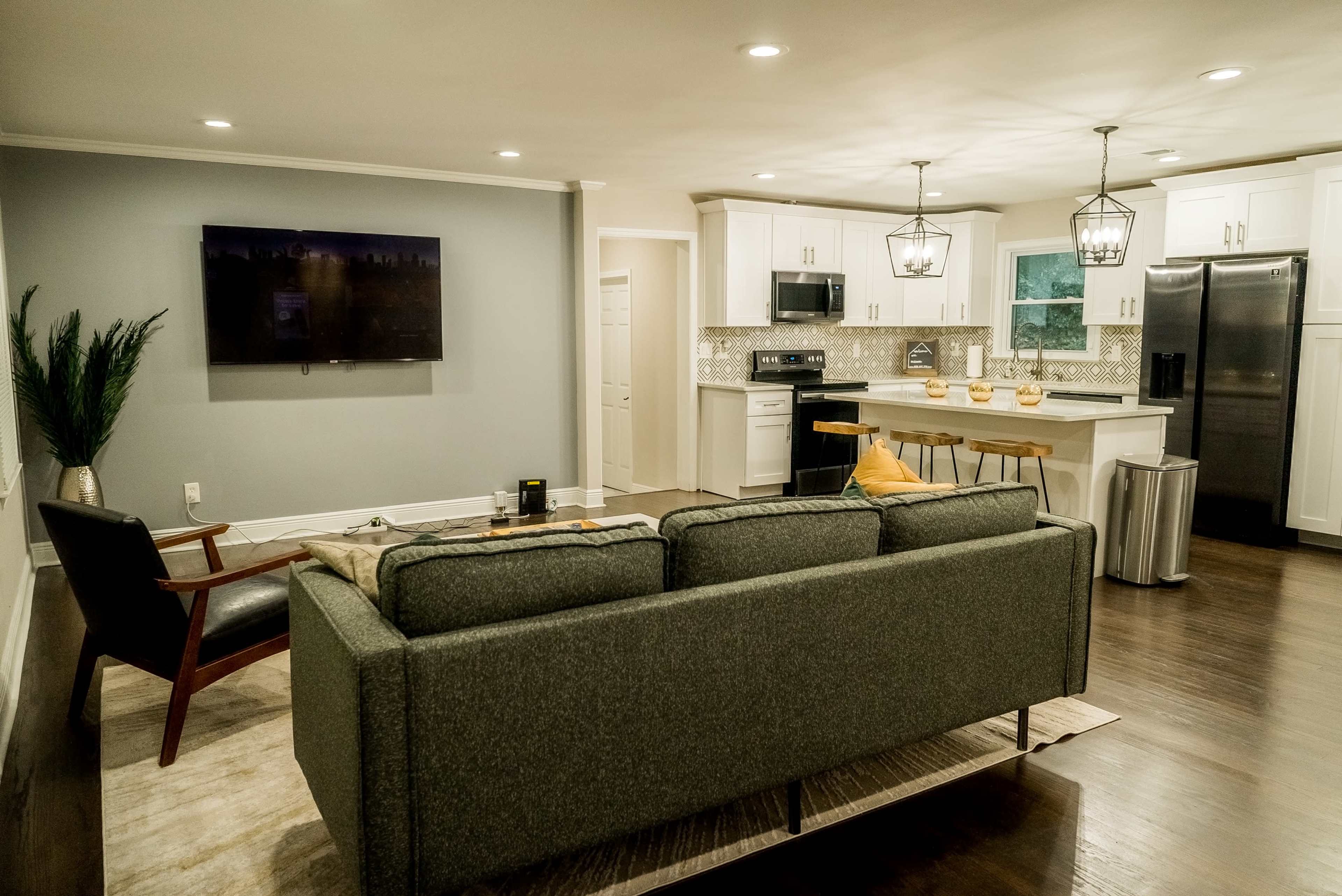 A modern living room that opens into a kitchen, featuring a gray sofa, a television on the wall, and white cabinetry.