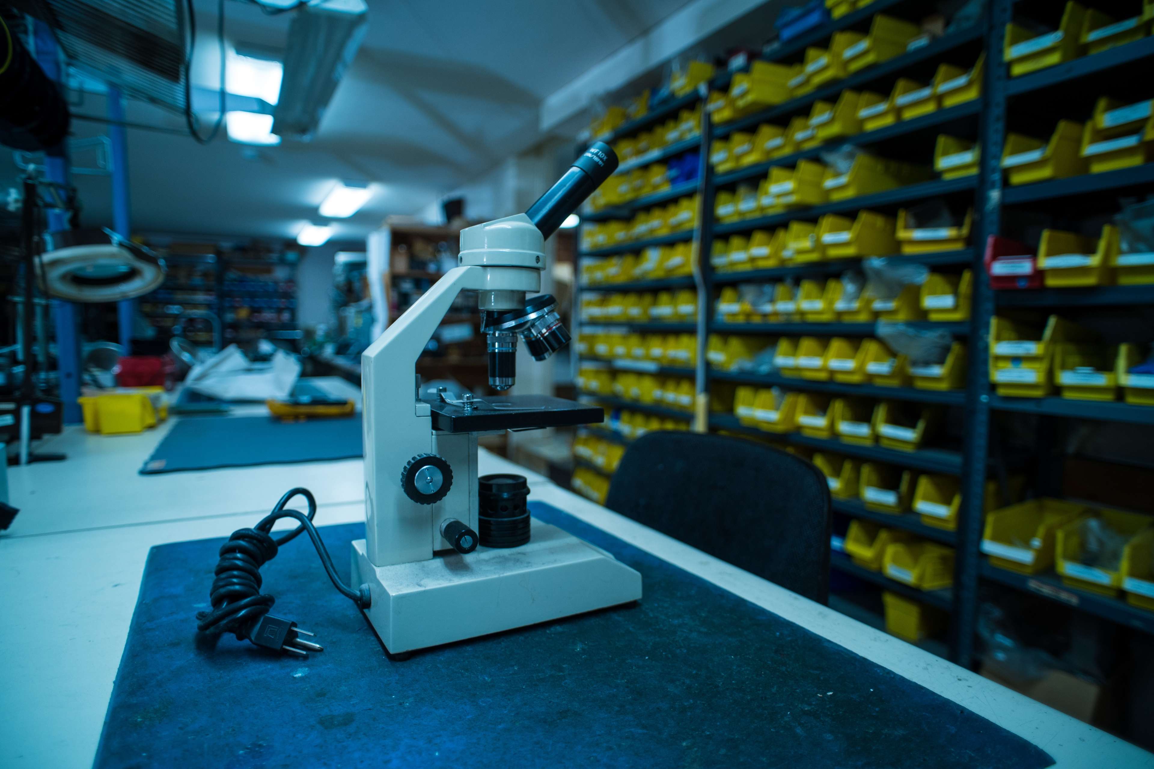 A microscope is positioned on a tabletop in a laboratory with shelves filled with yellow storage bins in the background.