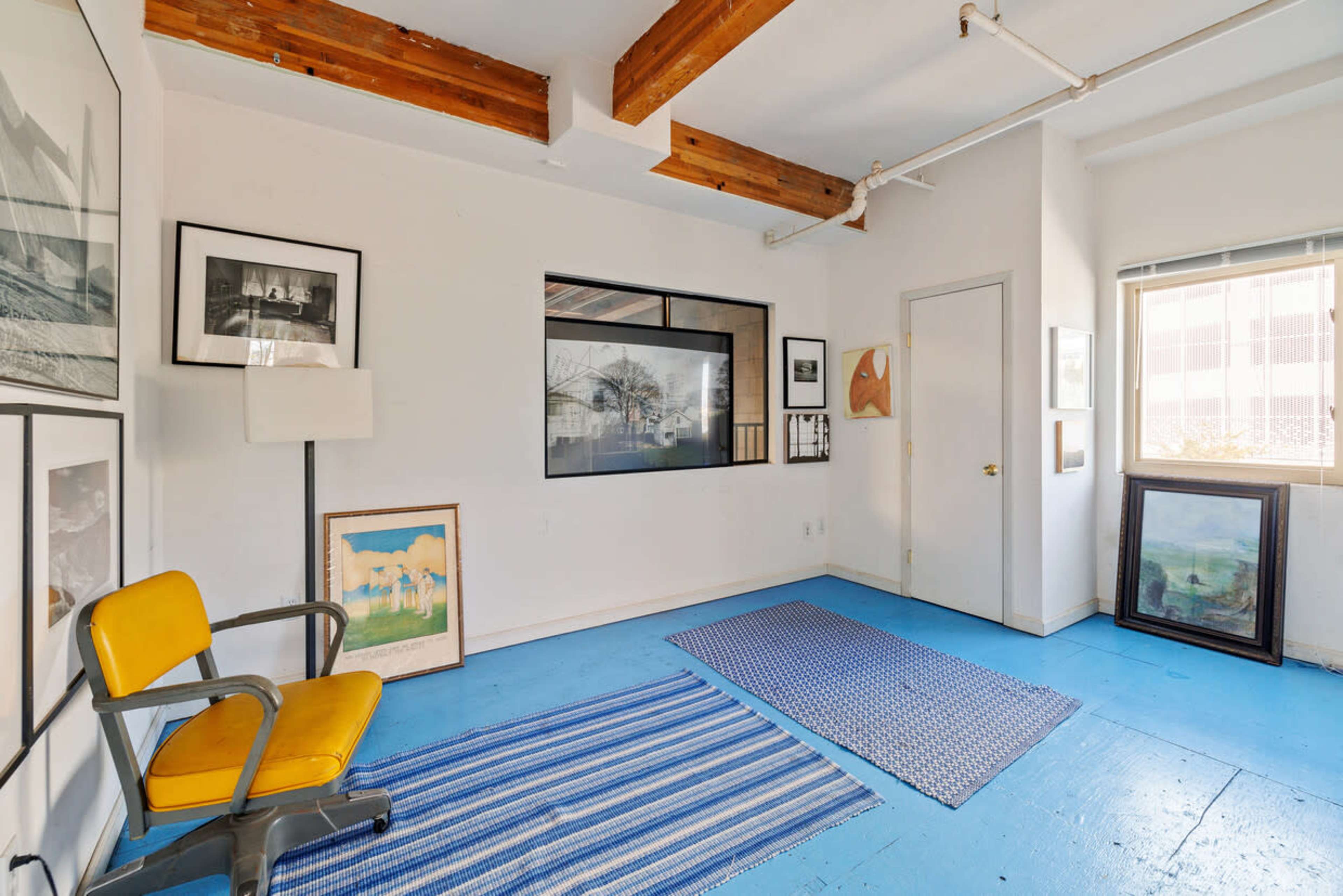 The room features blue flooring, wooden beams on the ceiling, a large window, and several artworks arranged on the walls and floor.