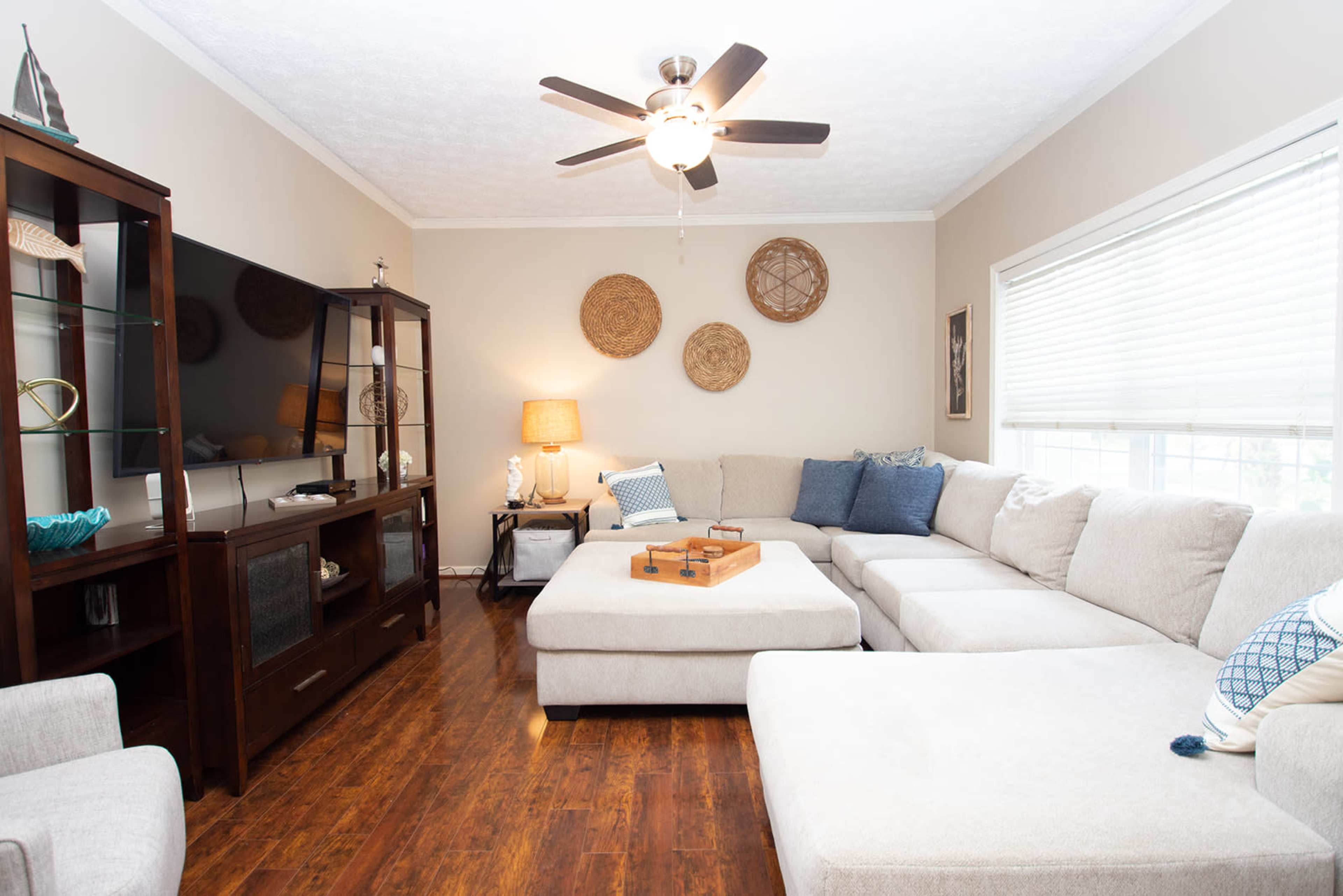 A large, light-colored sectional sofa is positioned in a cozy living room with hardwood floors, a flat-screen TV, and decorative wall hangings.