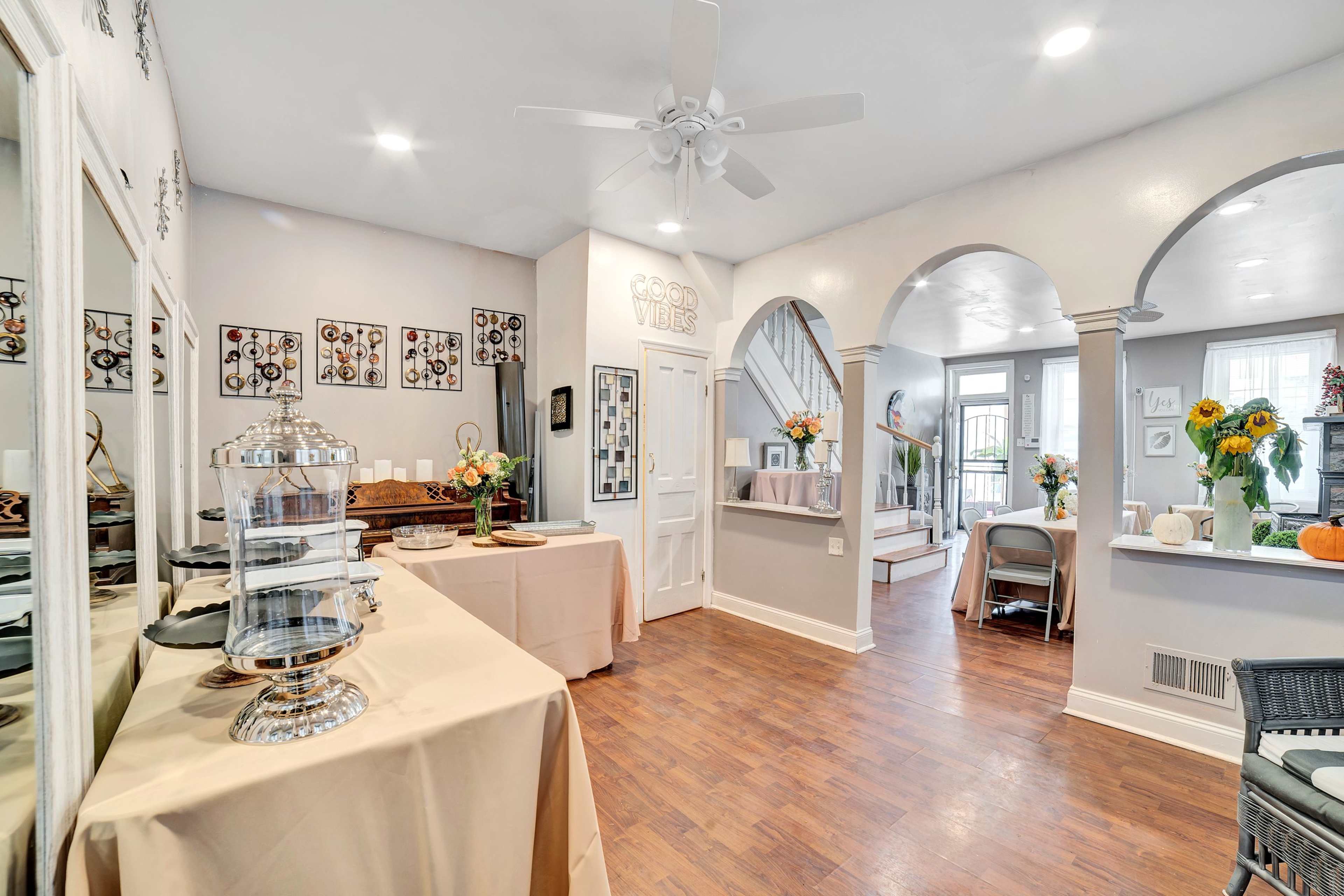 The image shows a well-lit room featuring a table with a decorative cover, a mirrored wall, and arches leading to another area, with flowers and a fan on the ceiling.