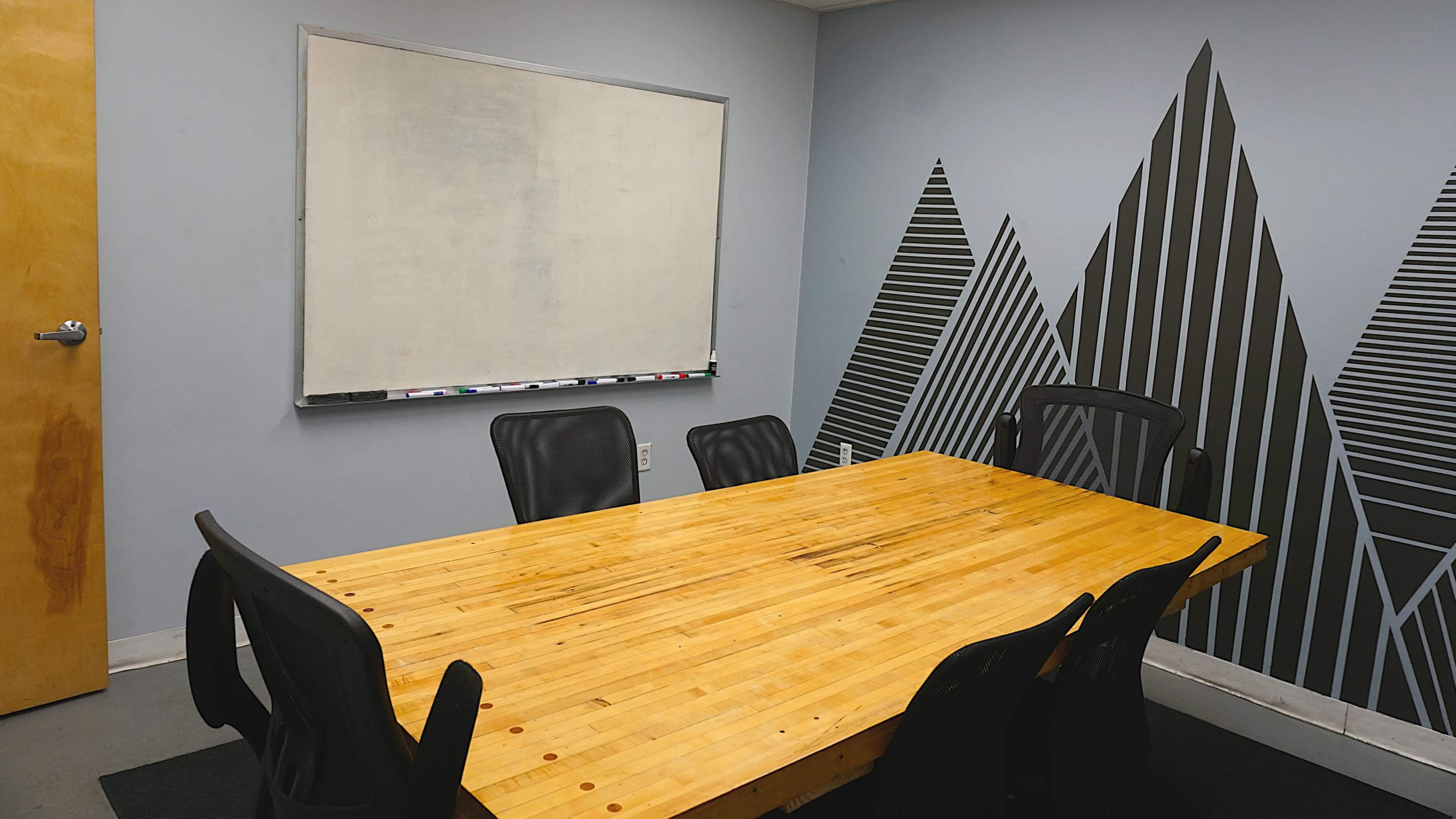 A conference room features a wooden table surrounded by black chairs, with a whiteboard and patterned wall decor in the background.