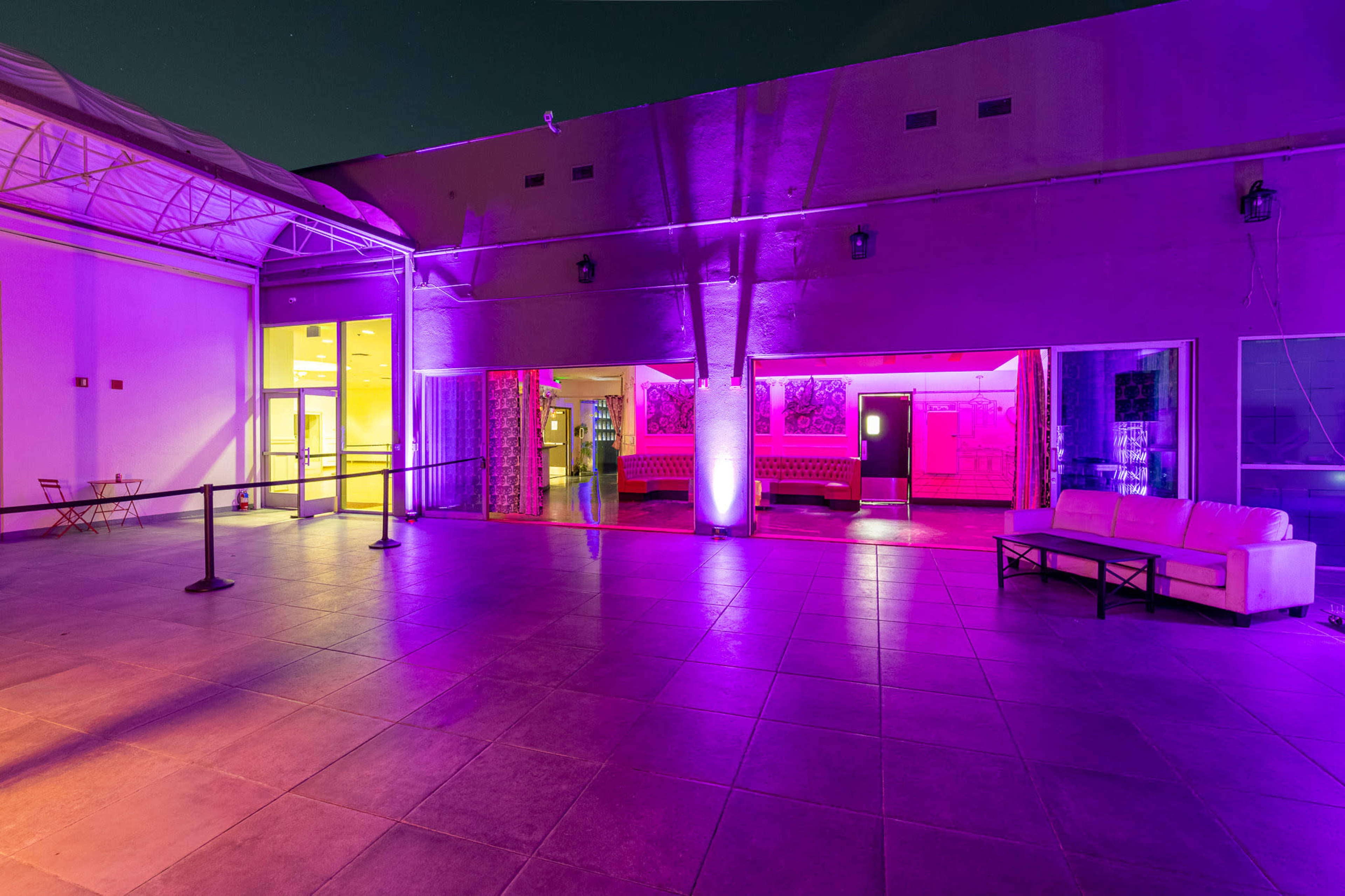 The image shows a spacious, illuminated outdoor area with purple lighting and modern furniture, leading into a building.