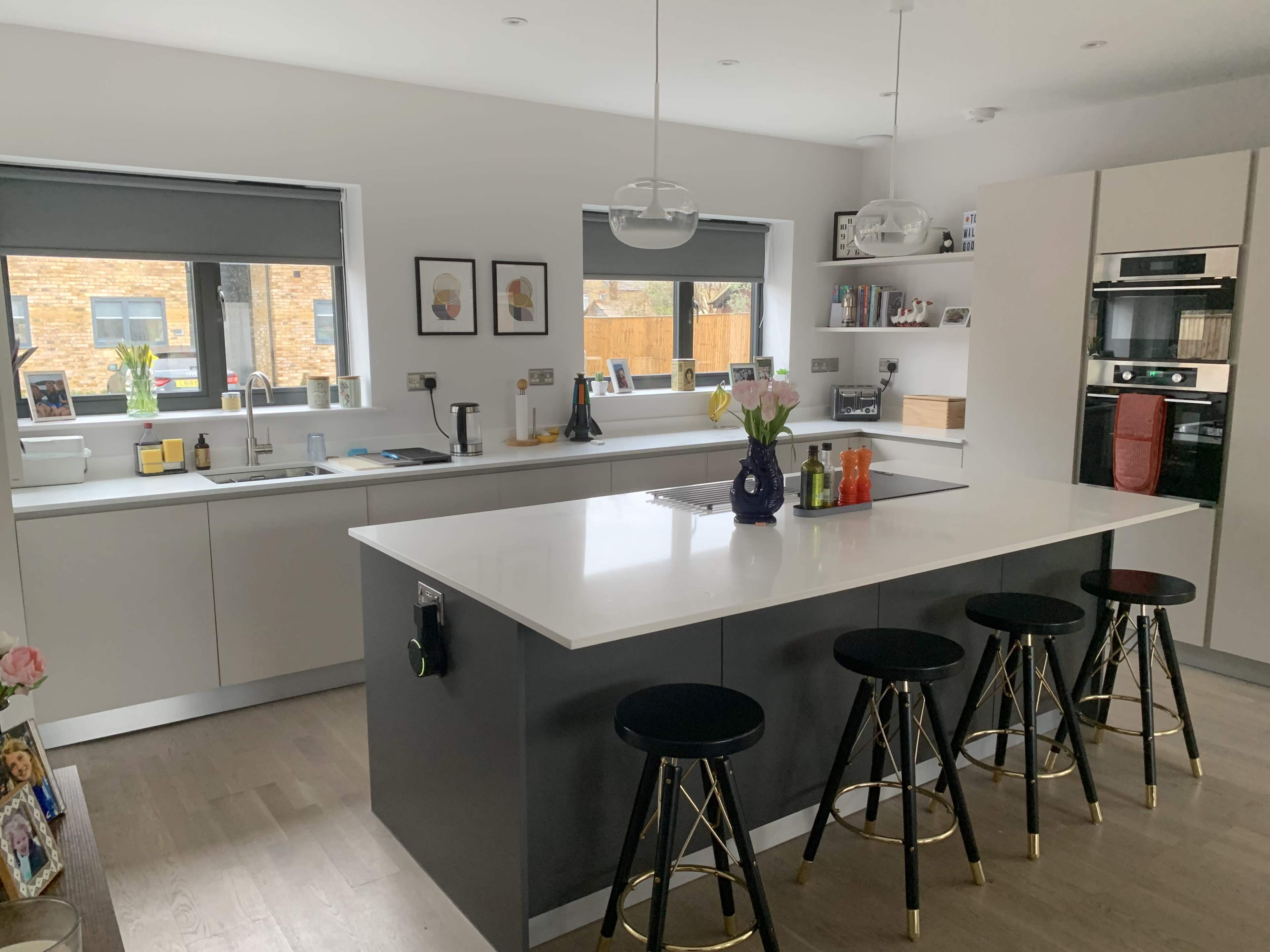 A modern kitchen features a large island with four black stools, white countertops, and sleek cabinetry, along with framed artwork and a window view.