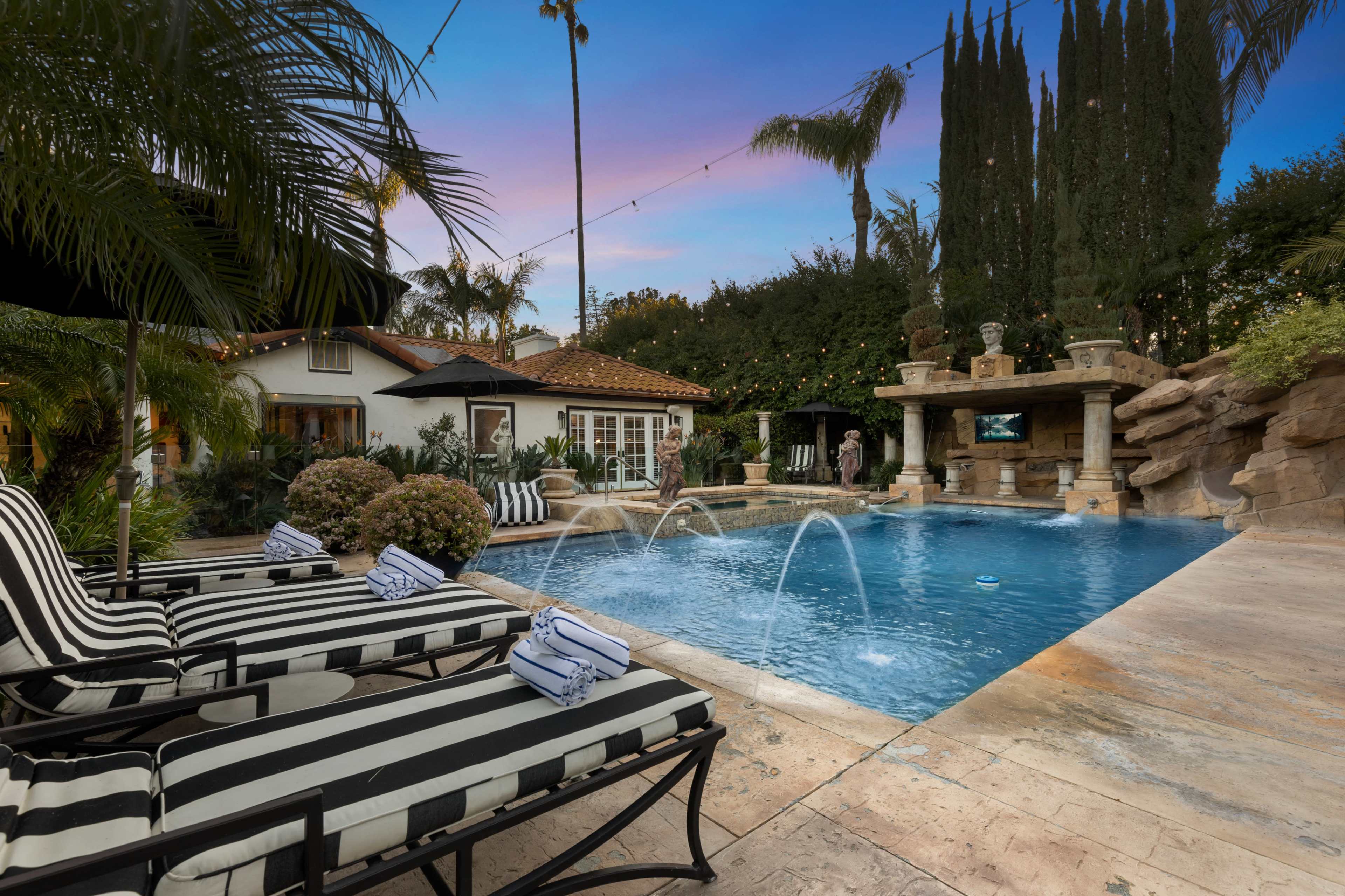 A pool with water features is surrounded by lounge chairs and lush landscaping, with a residential building and stone structure in the background.