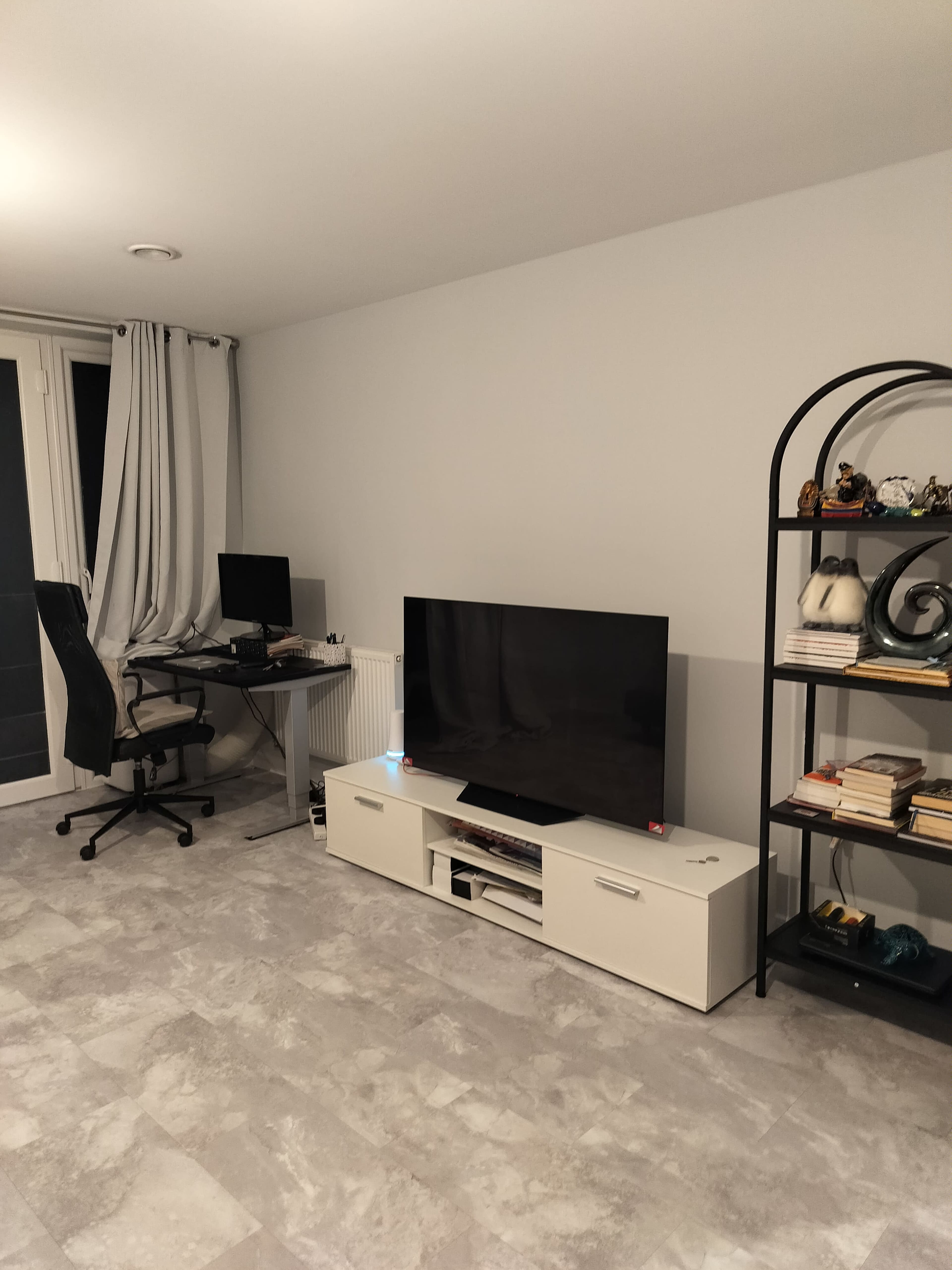 The image shows a modern living room with a television on a white media console, a black bookshelf filled with books and decor, and a desk with a computer in the corner.