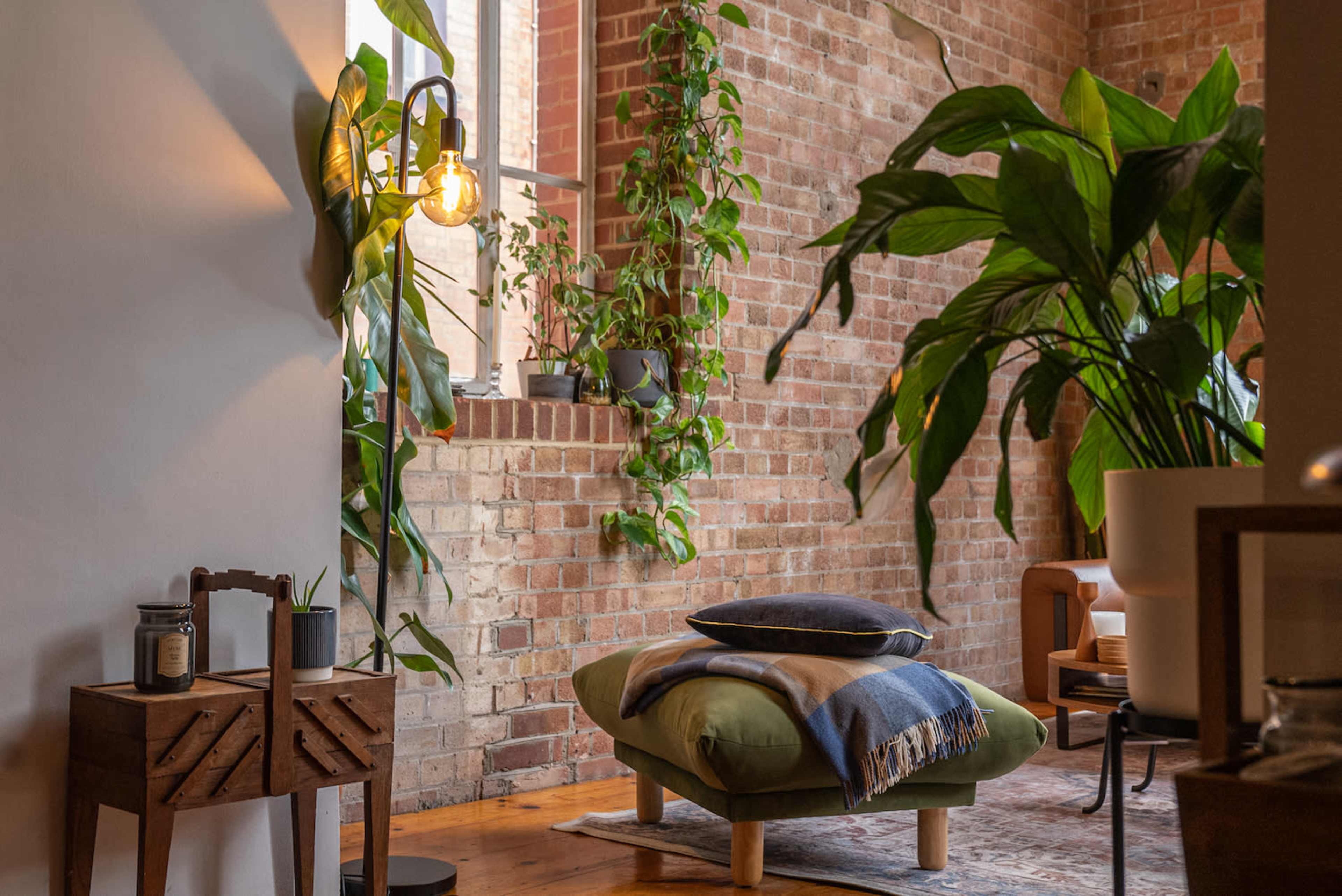 A cozy interior with a green ottoman, potted plants, a wooden side table, and exposed brick walls.