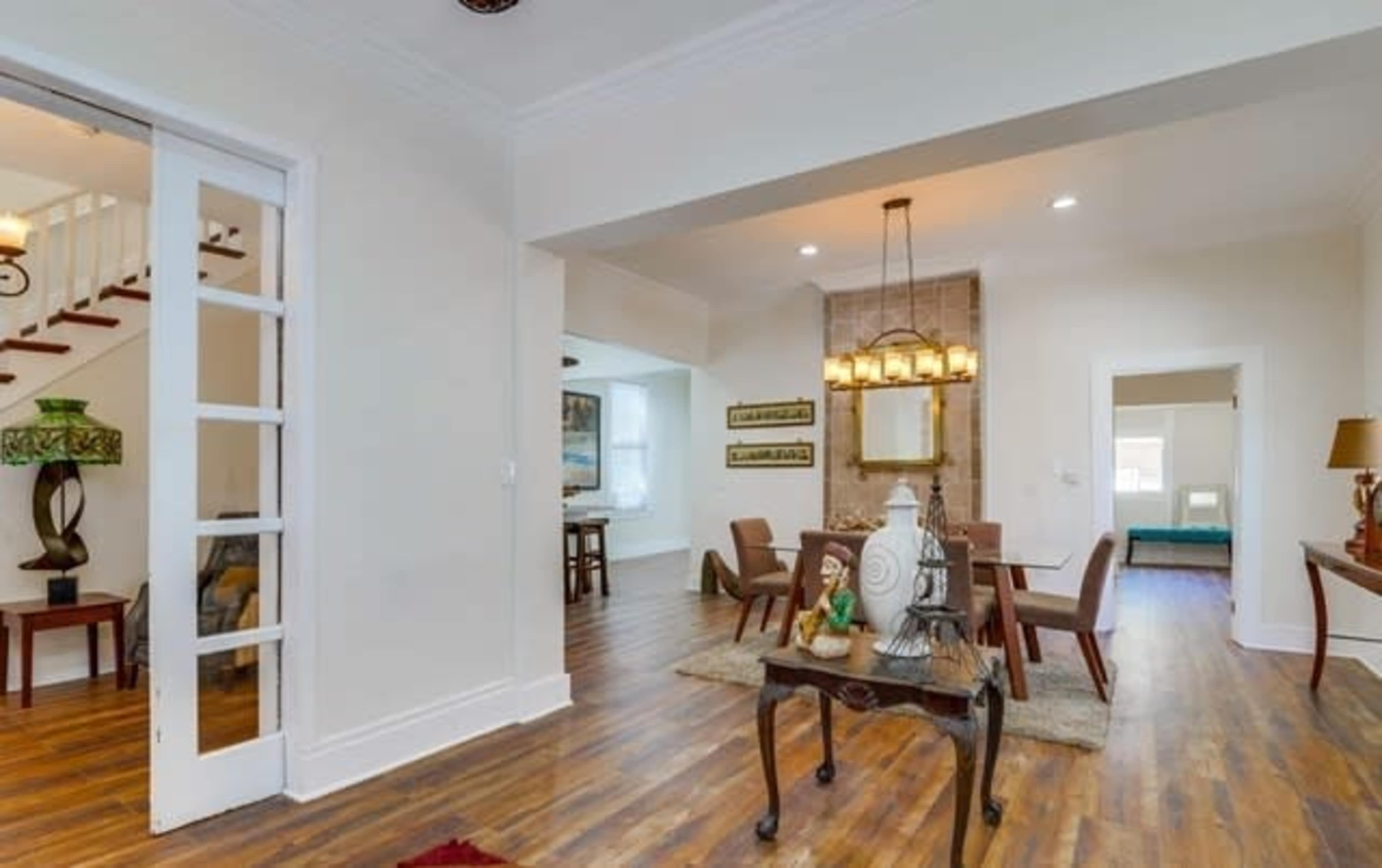 A light-filled dining area with a central pendant light, wooden flooring, and a glimpse of a staircase in the background.