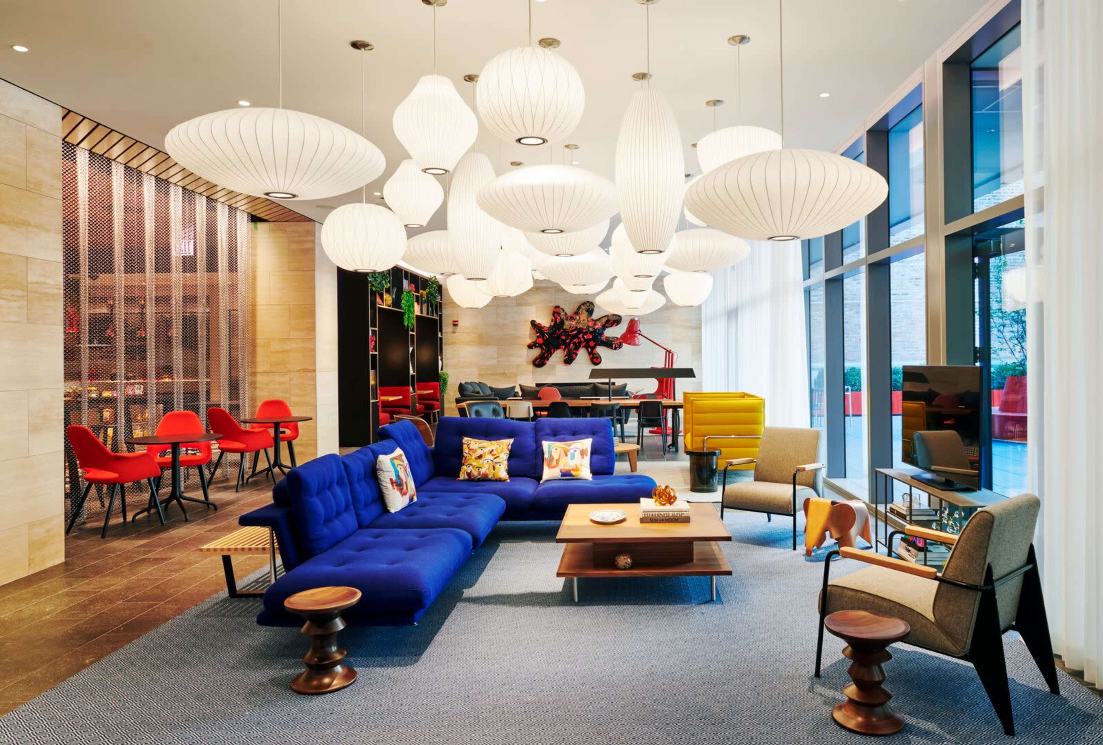 The scene features a modern lounge area with a blue sectional sofa, colorful chairs, unique pendant lights, and a large window allowing natural light to fill the space.