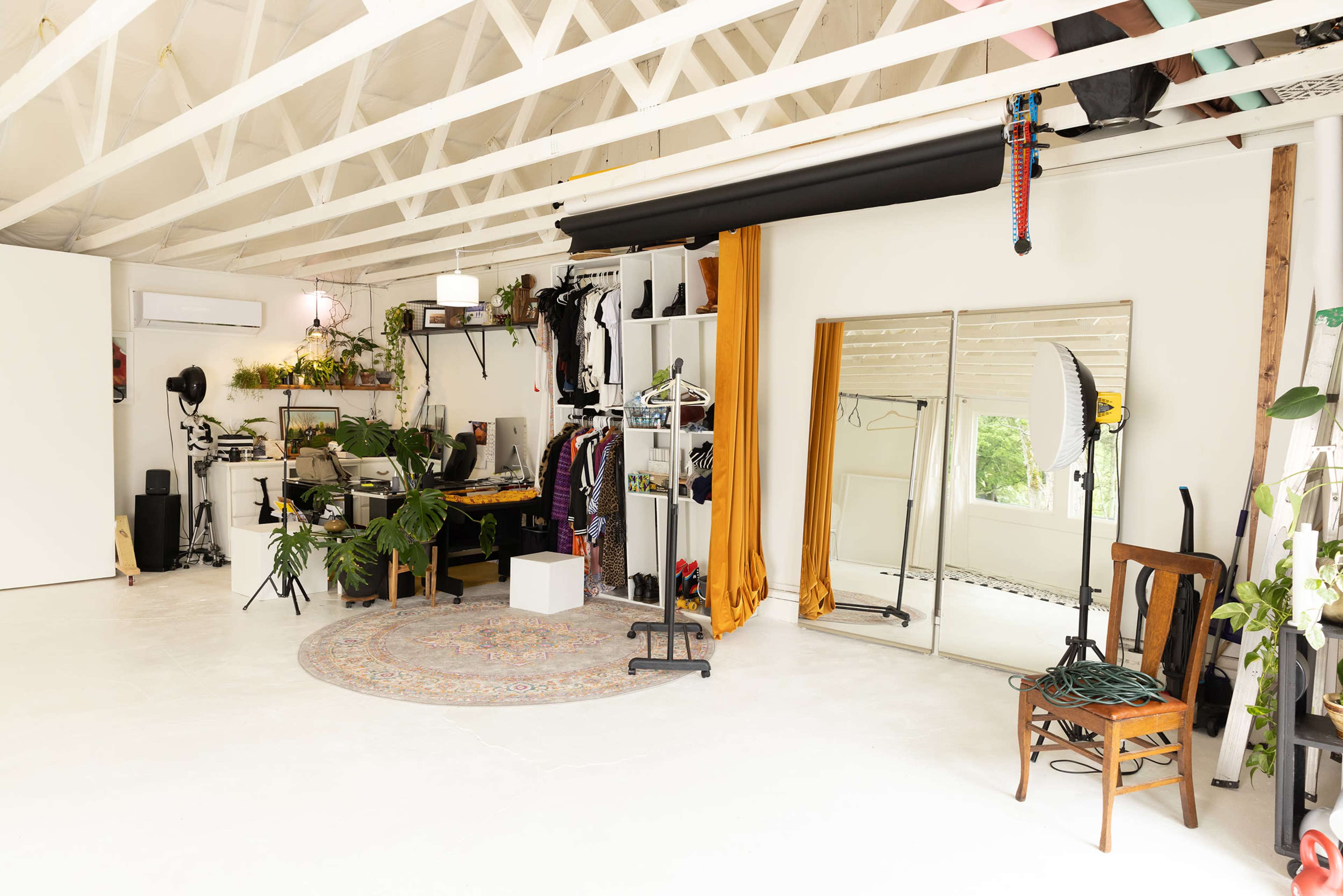 The image shows a spacious studio with a white floor, large mirrors, clothing racks, indoor plants, and a bright work area.