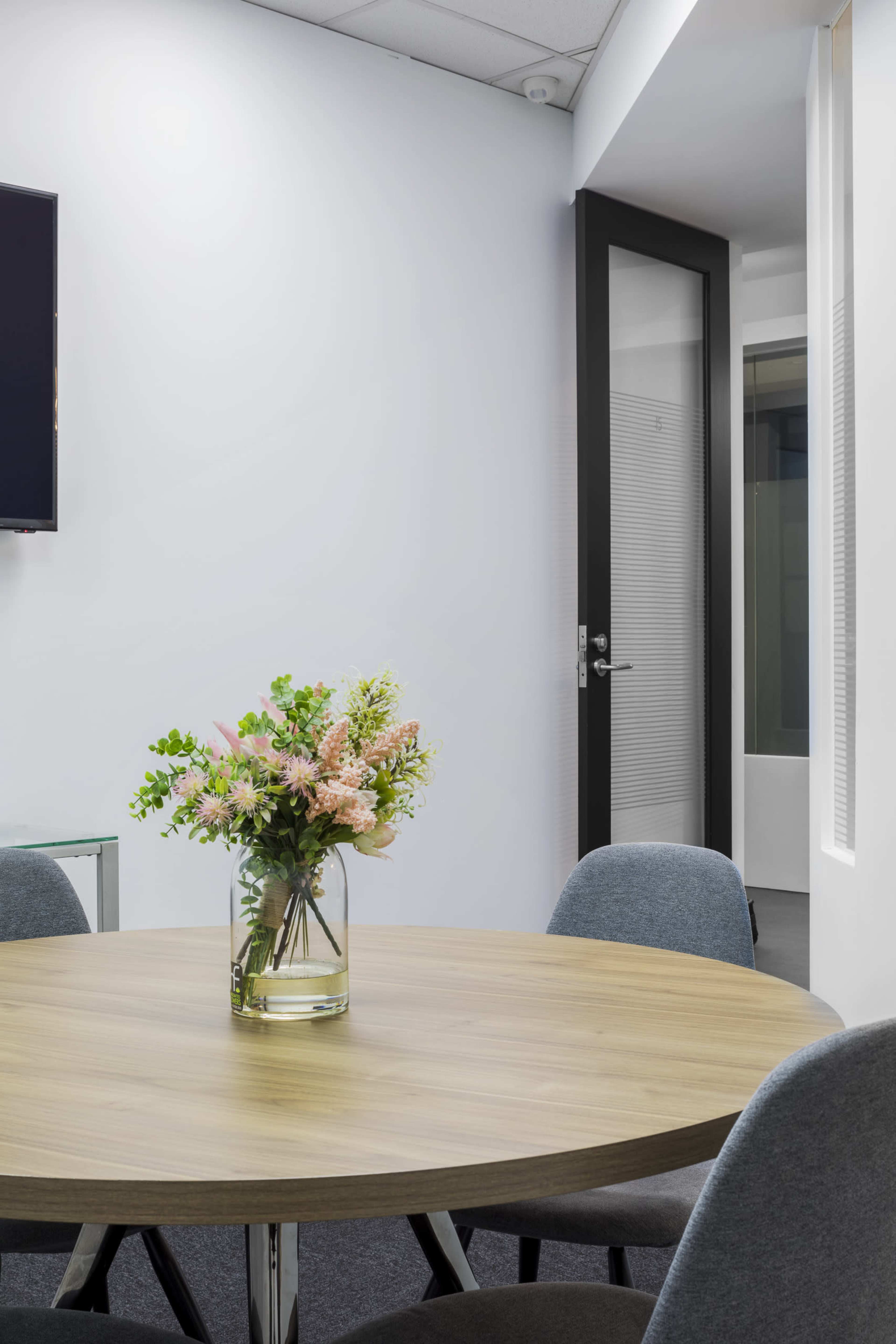 A round wooden table with a vase of flowers sits in the foreground, while a glass door leads to another room in the background.