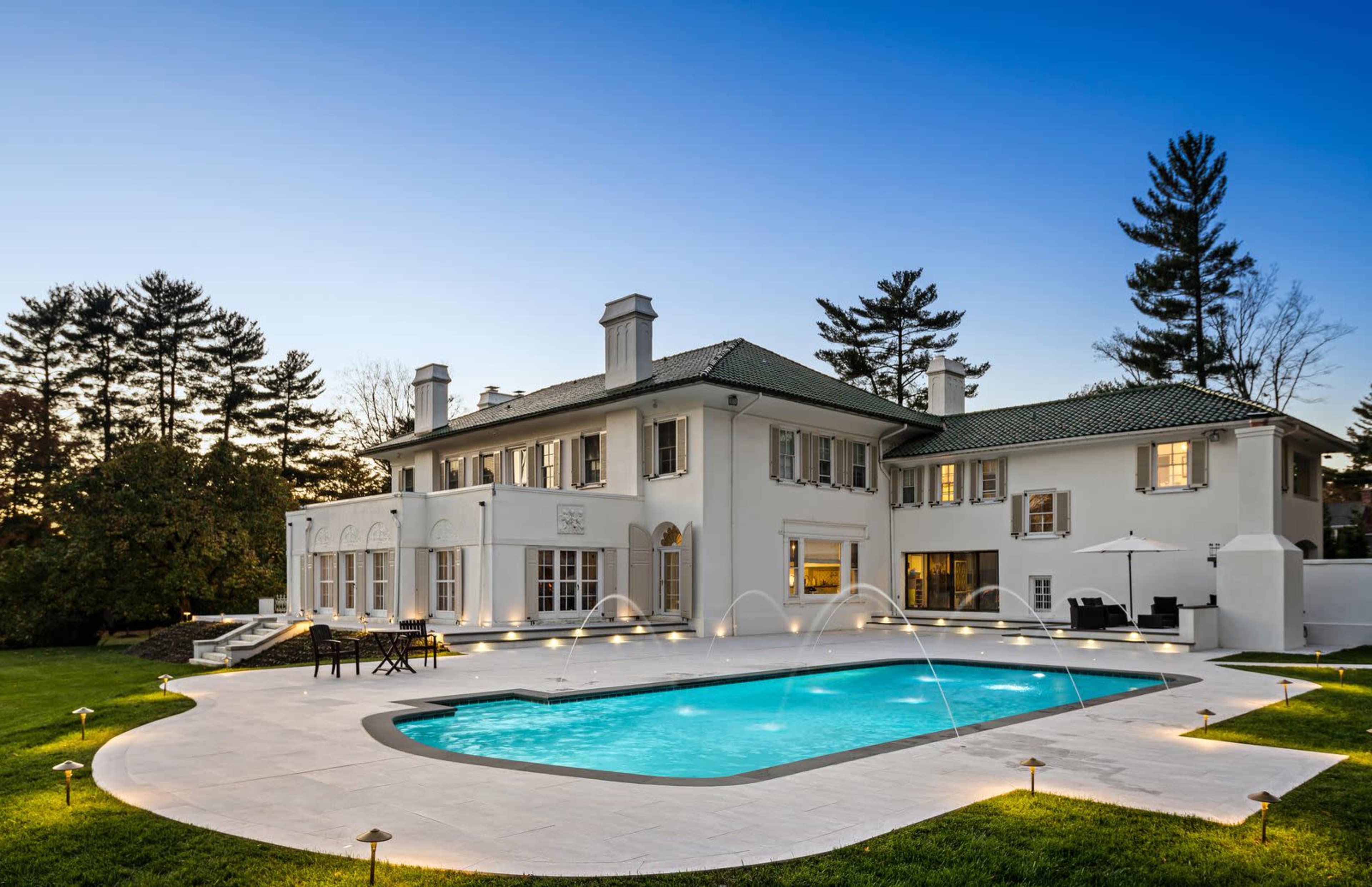 The image shows a modern white mansion with a large swimming pool and landscaped yard, surrounded by trees at twilight.