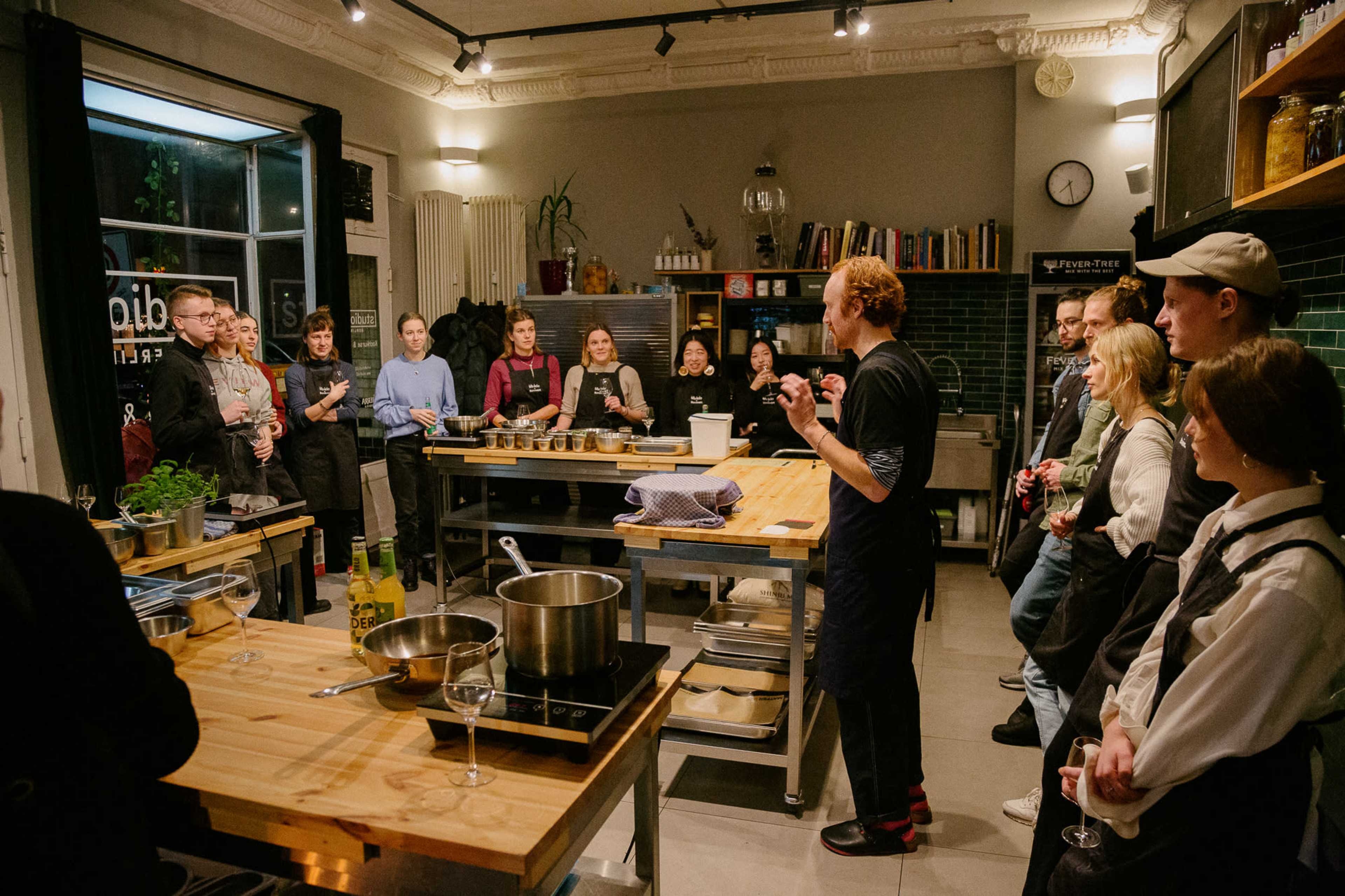 A group of people gathers in a modern kitchen setting as a man speaks to the audience while demonstrating a cooking technique.
