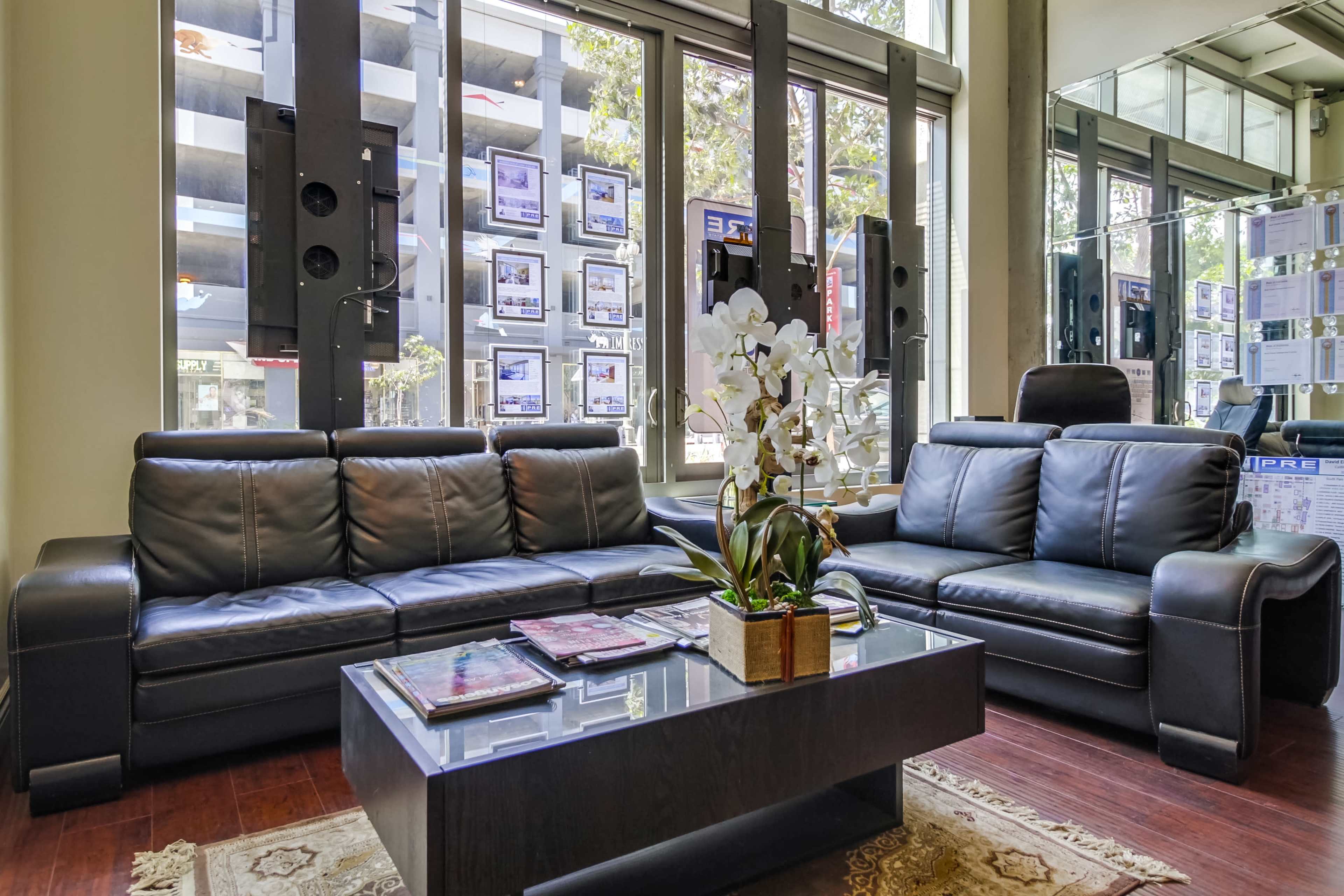 The image shows a modern seating area with two black leather sofas facing a glass coffee table, surrounded by large windows displaying various real estate listings.
