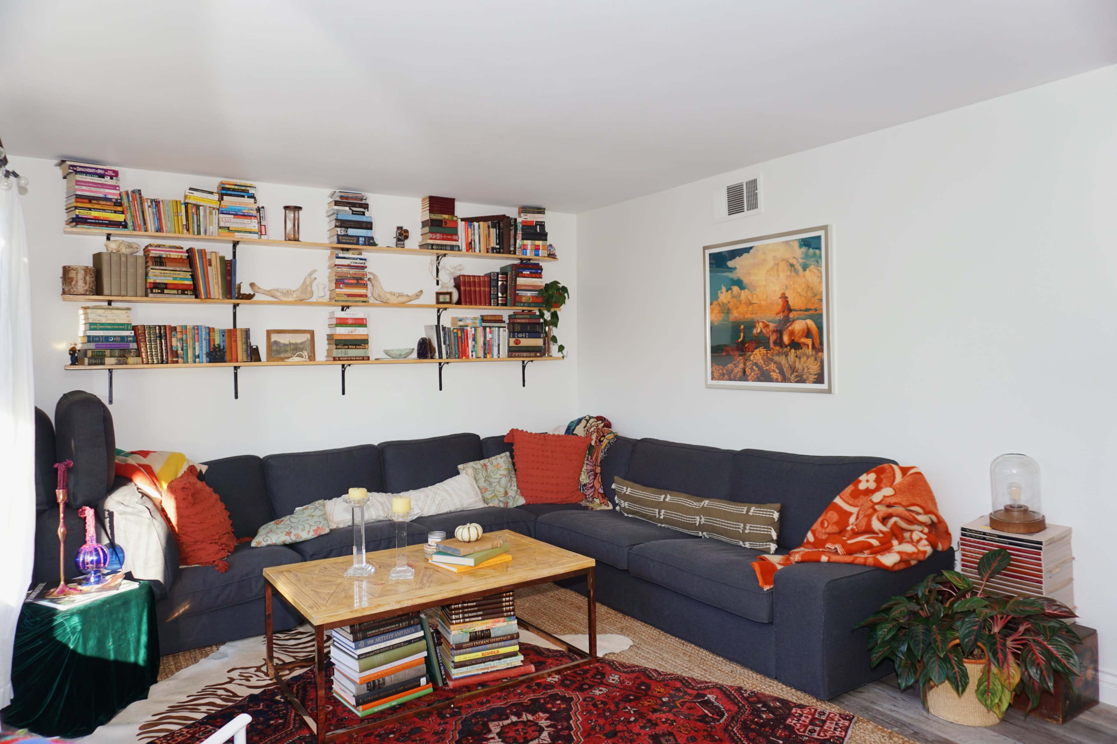 A cozy living room features a dark sectional sofa, a wooden coffee table with books stacked atop it, and shelves filled with books along the wall.