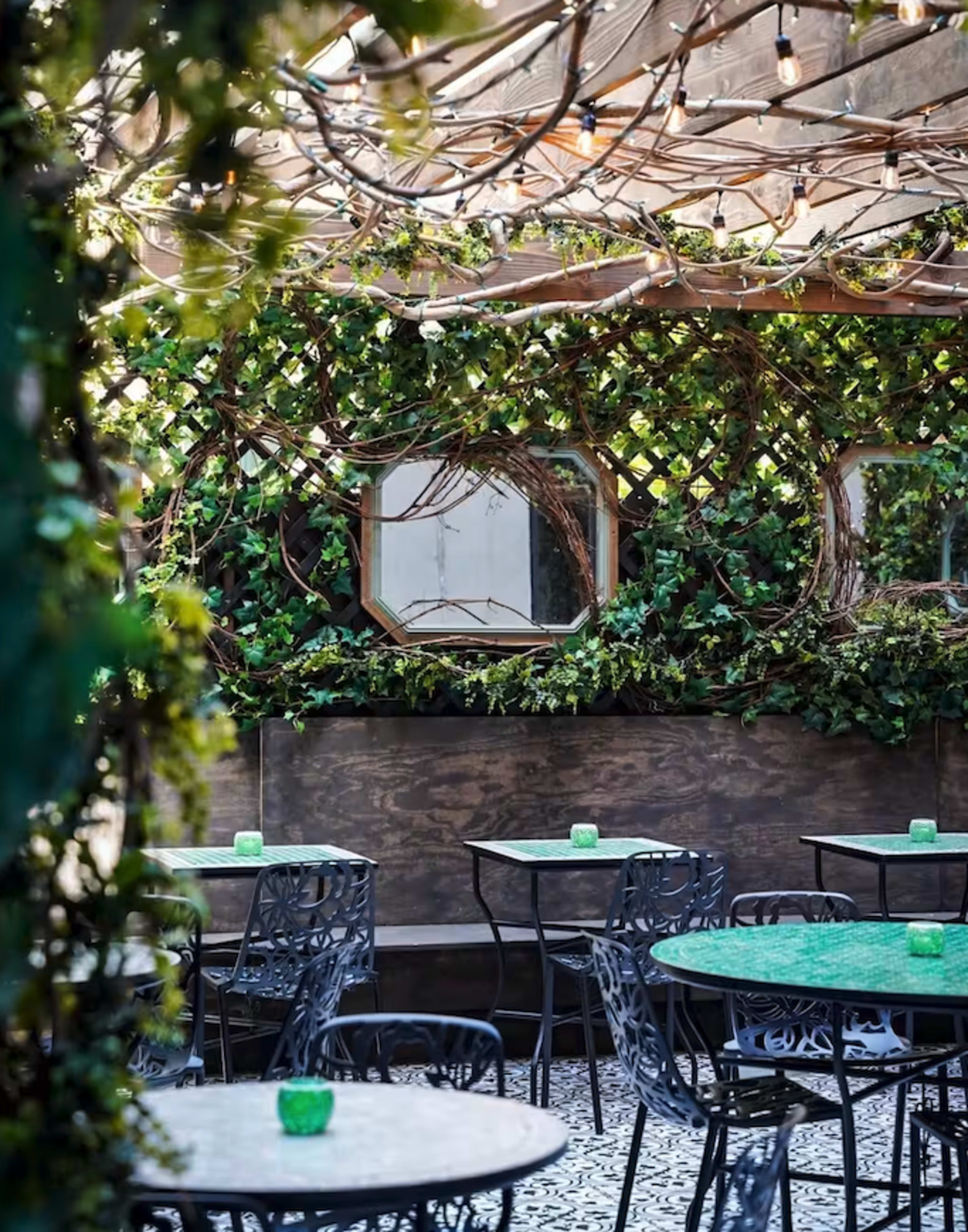 The image shows a cozy outdoor dining area adorned with ivy-covered walls, circular mirrors, and small round tables.