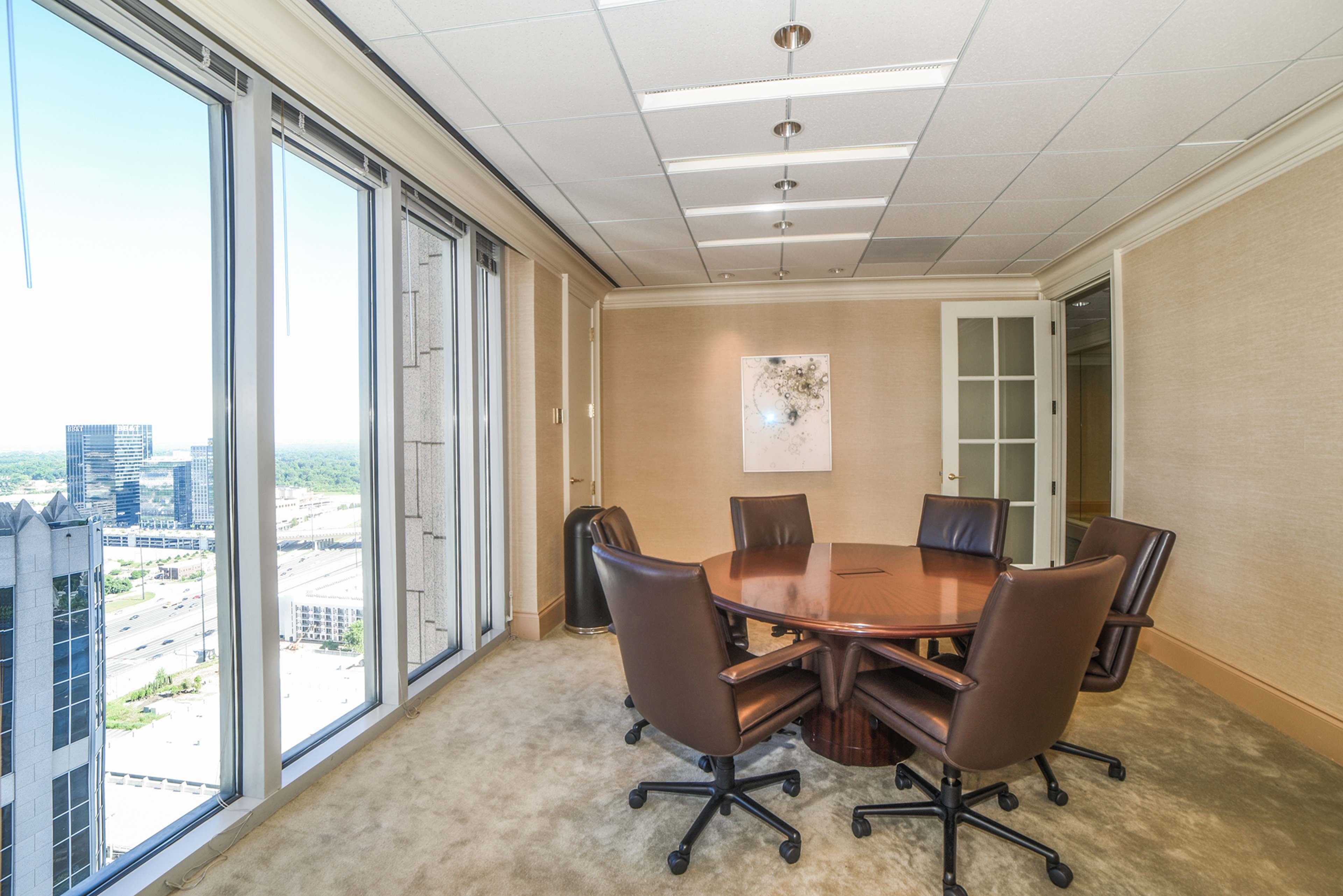 A conference room with a wooden round table surrounded by six brown chairs, featuring large windows that offer a view of the city.