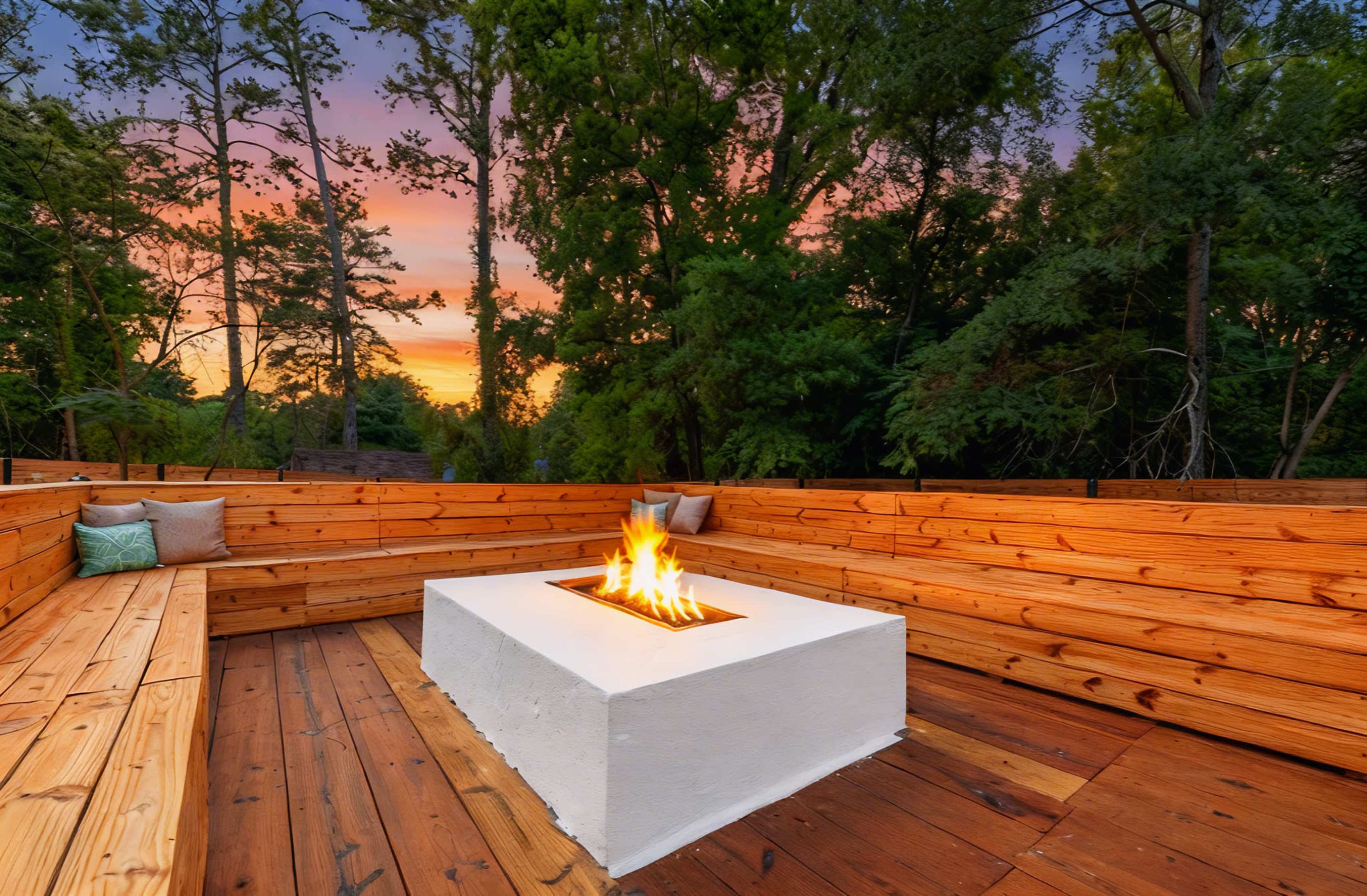 A cozy outdoor seating area features wooden benches surrounding a rectangular fire pit, with a vibrant sunset and trees in the background.