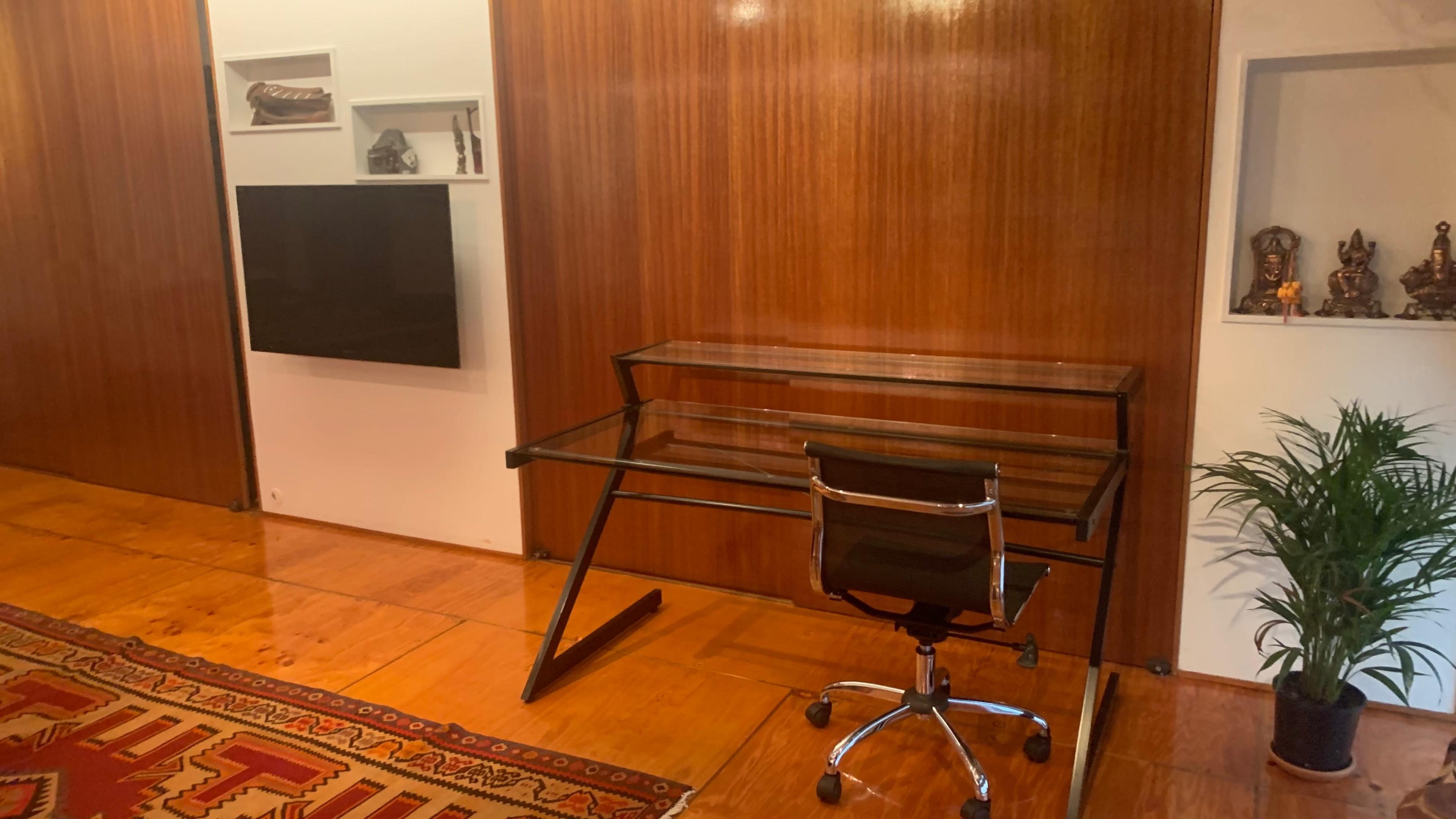 A minimalist office space featuring a glass desk with a rolling chair against a wooden wall, a television mounted on the wall, and decorative items on shelves.