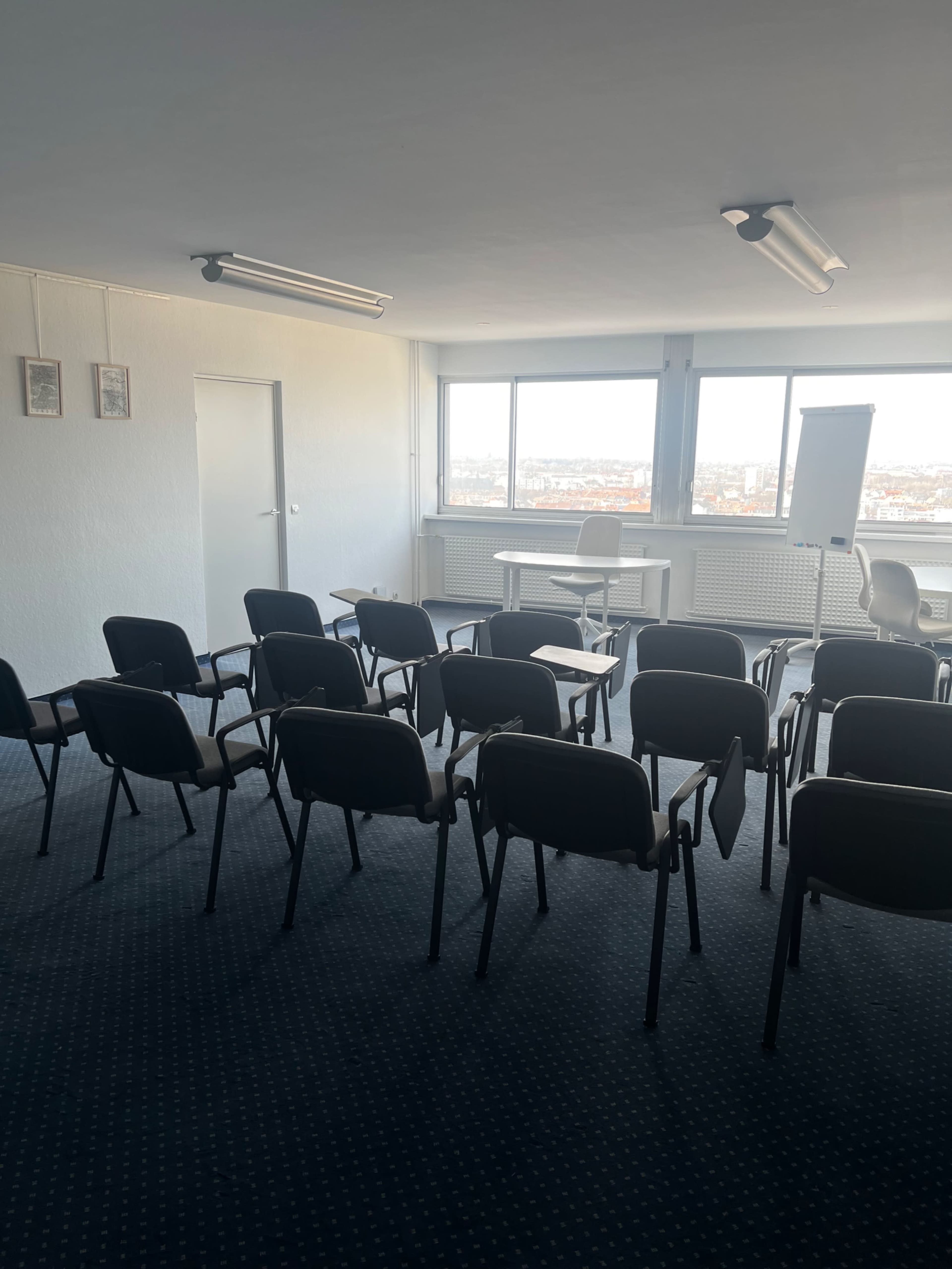 A spacious room is set up with rows of chairs facing a table and a flip chart, with large windows allowing natural light in.