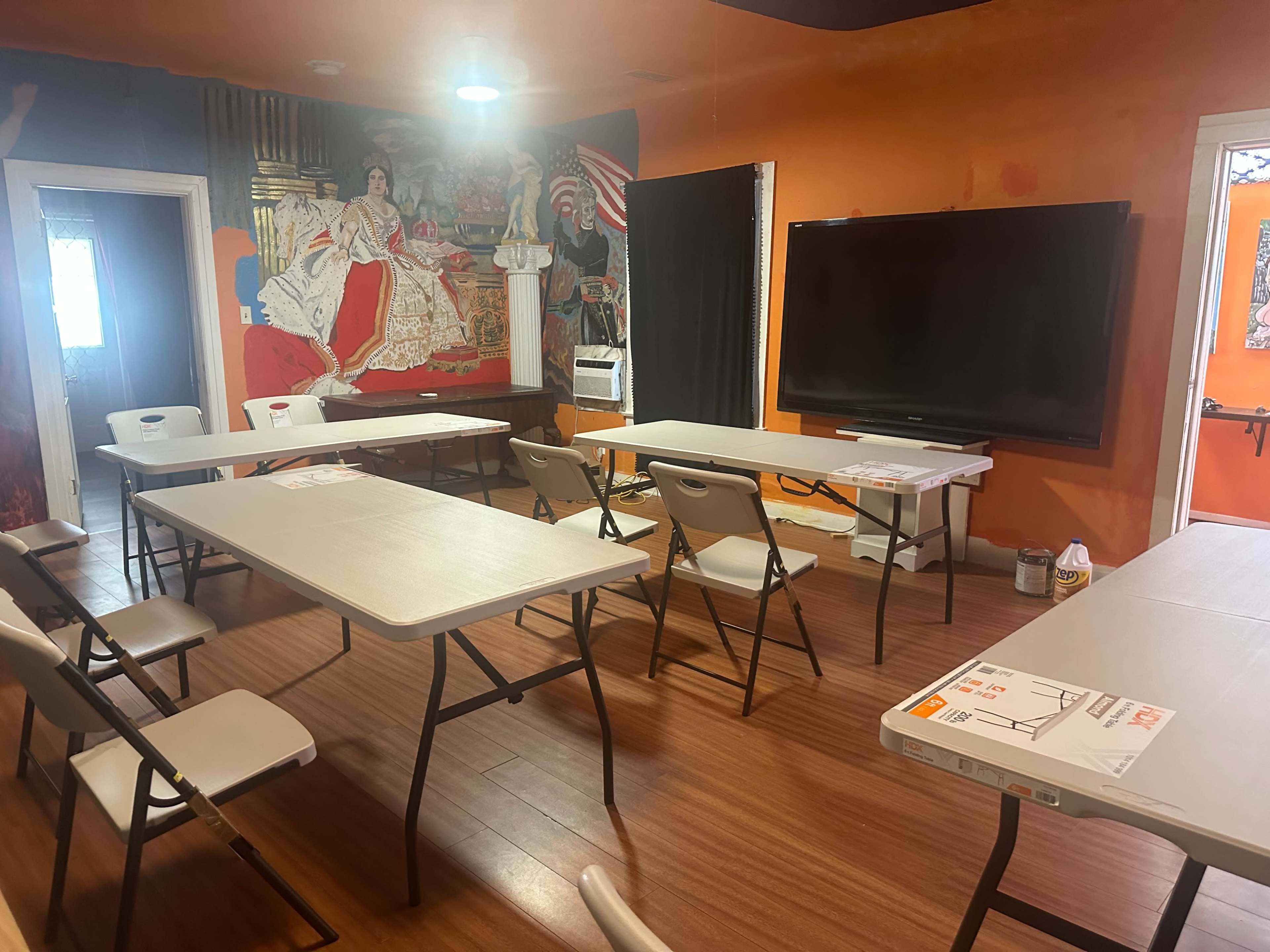 The image shows a meeting room with several folding tables arranged in a U-shape, equipped with chairs and a large television mounted on the wall, featuring a mural in the background.