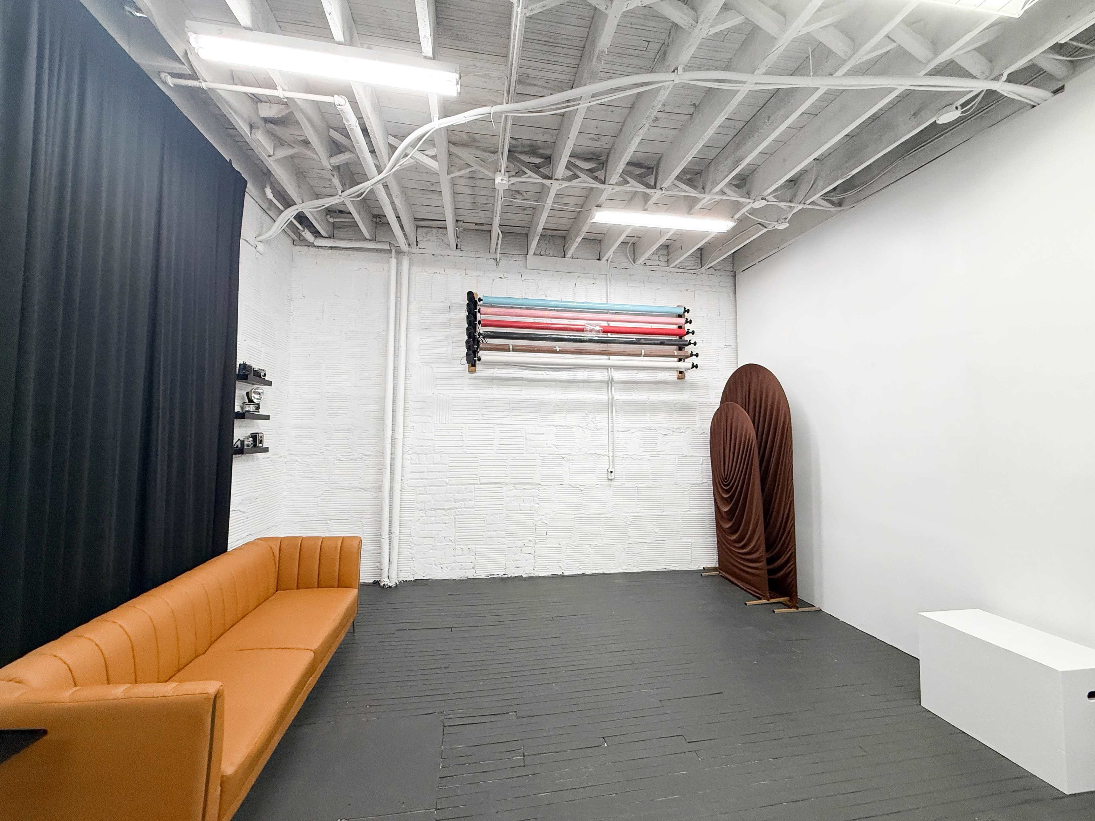 The image shows a minimalist studio space with a brown sofa, a roll of backdrop materials on a wall, and a sculptural element on the right.